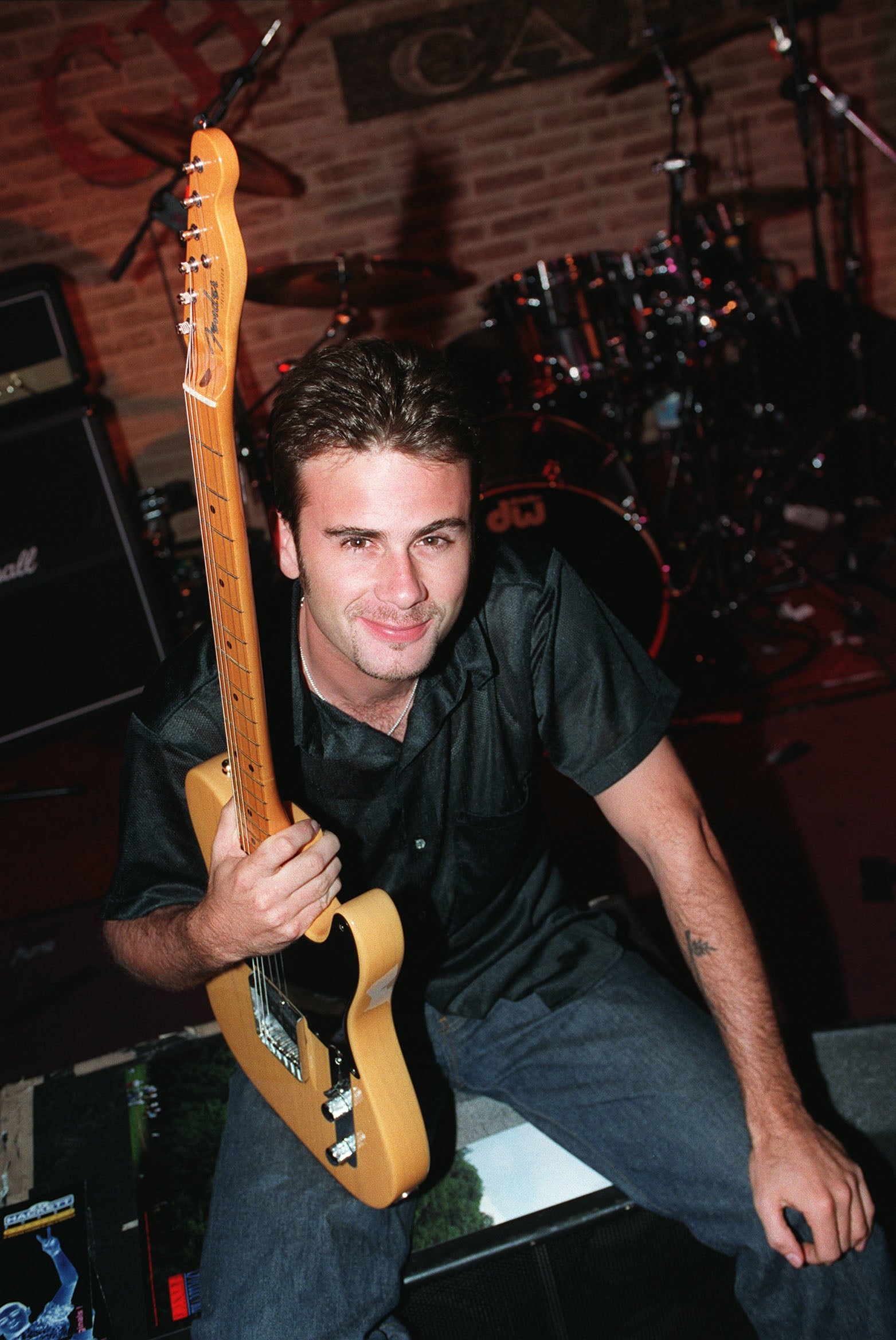 Person holding a guitar, sitting on stage with musical equipment in the background. Casual clothing style