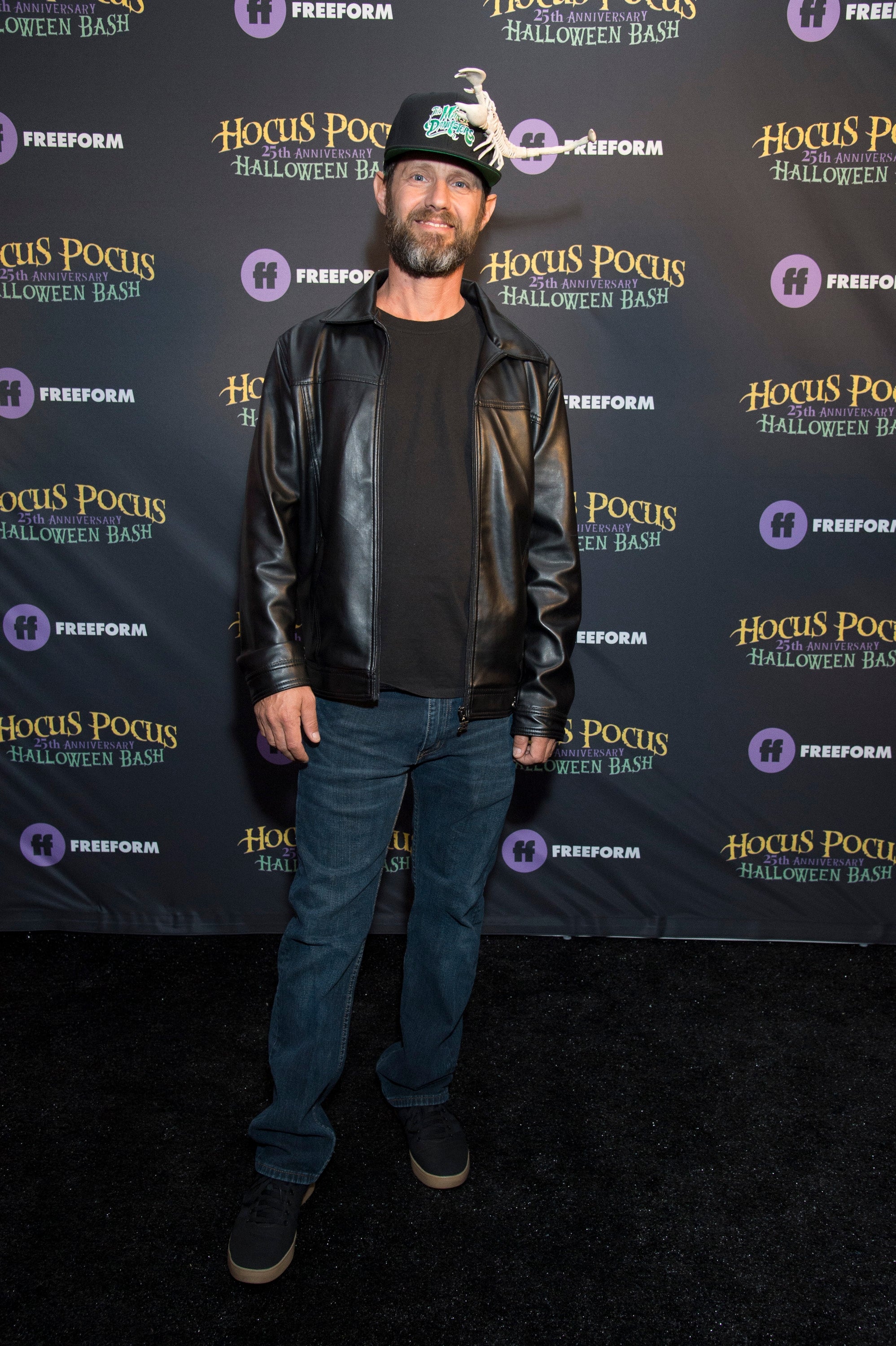 A person on a red carpet wearing a leather jacket, jeans, and a cap at the "Hocus Pocus Halloween Bash" event