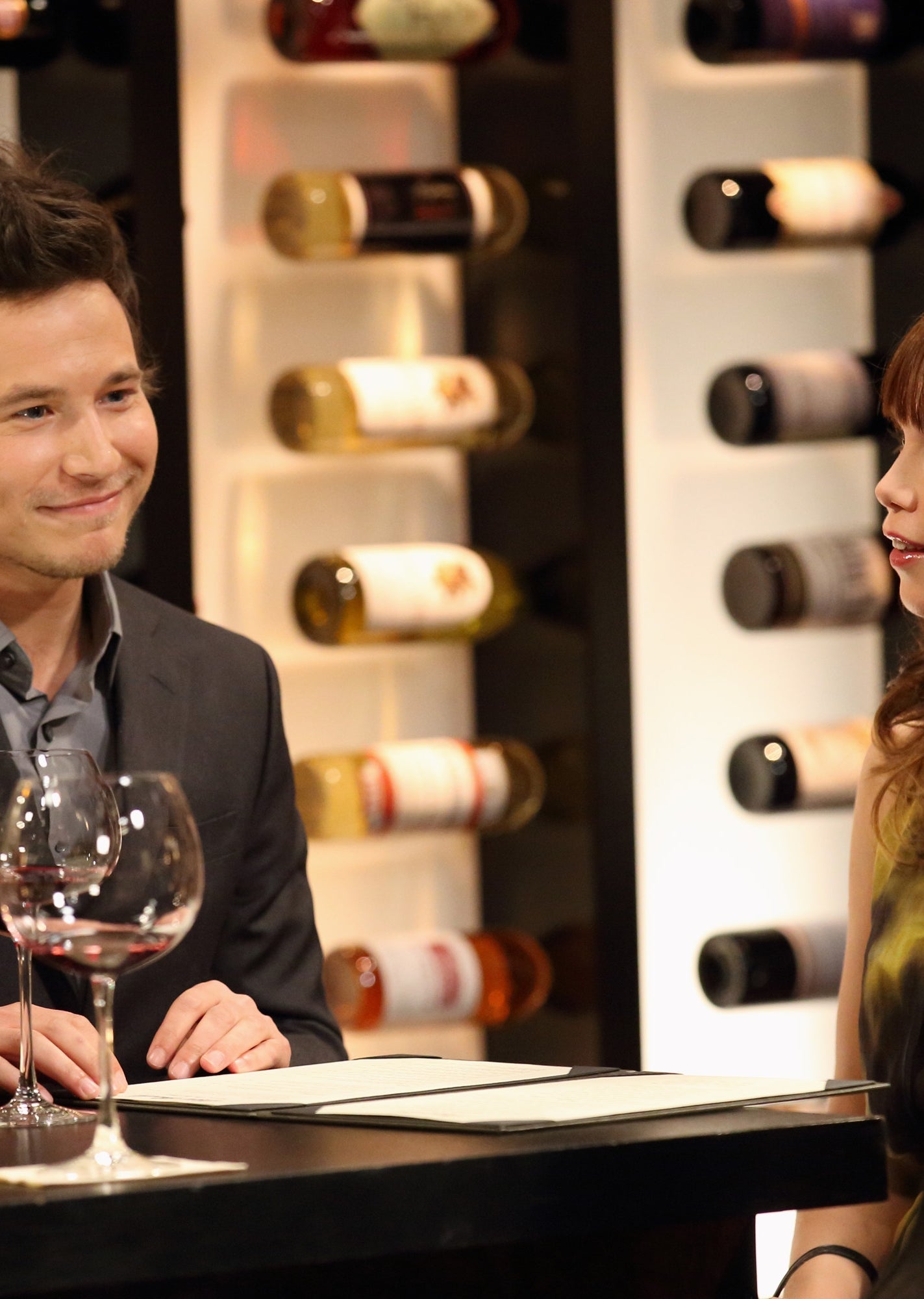 A group of people are sitting at a dimly-lit wine bar, chatting and smiling, with bottles of wine on display in the background