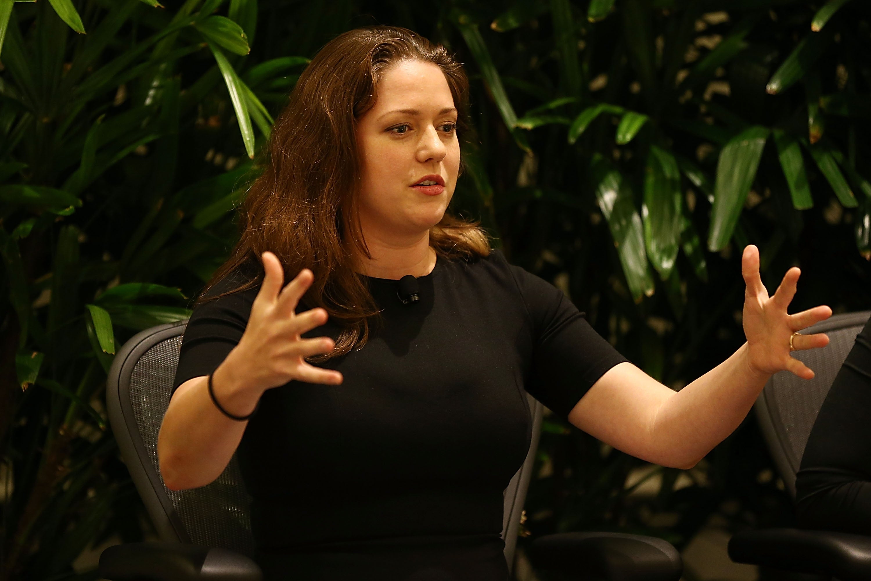 A woman gestures while speaking, seated against a backdrop of leafy plants, wearing a simple, elegant black dress