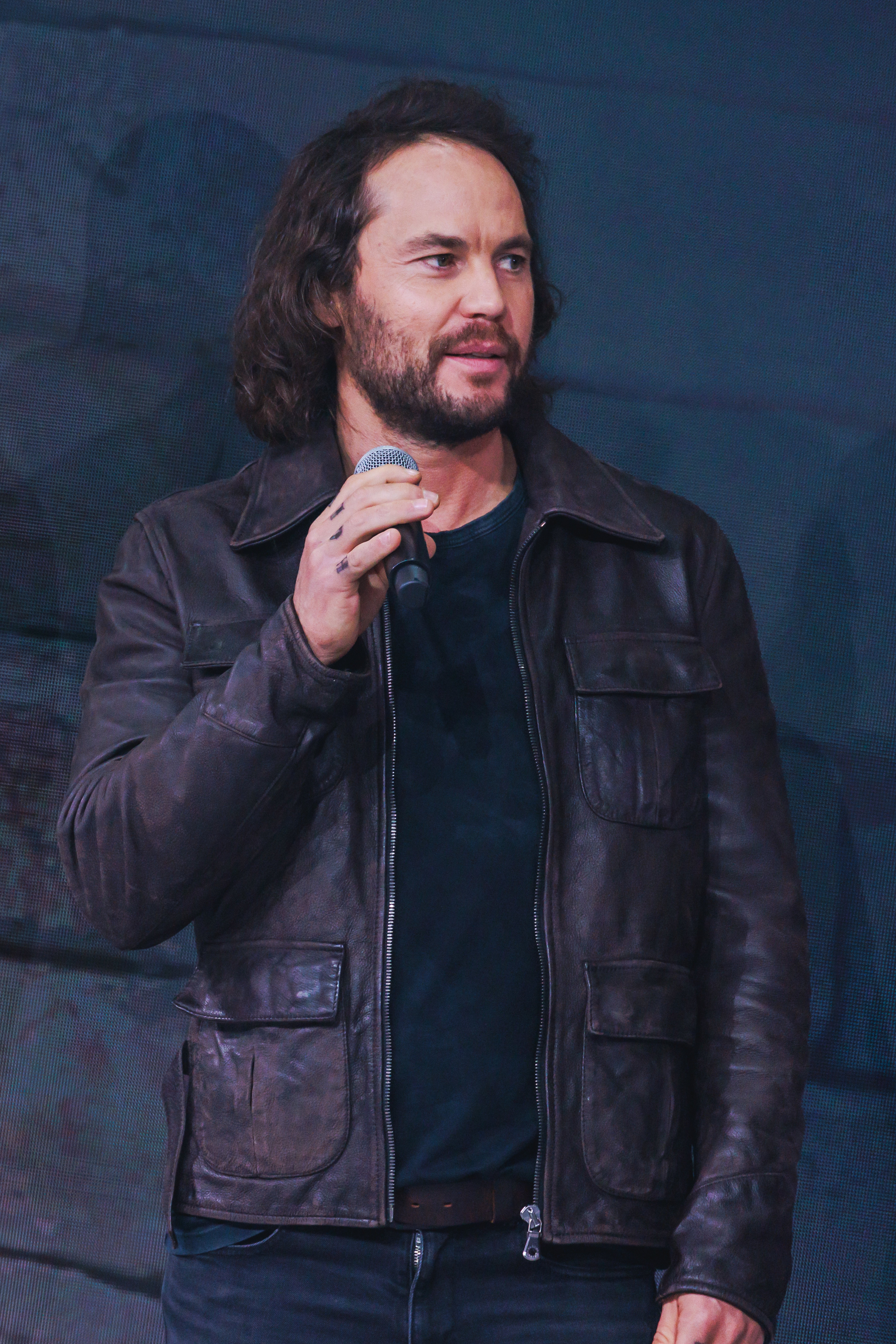 A person on stage holds a microphone, wearing a casual leather jacket over a dark shirt