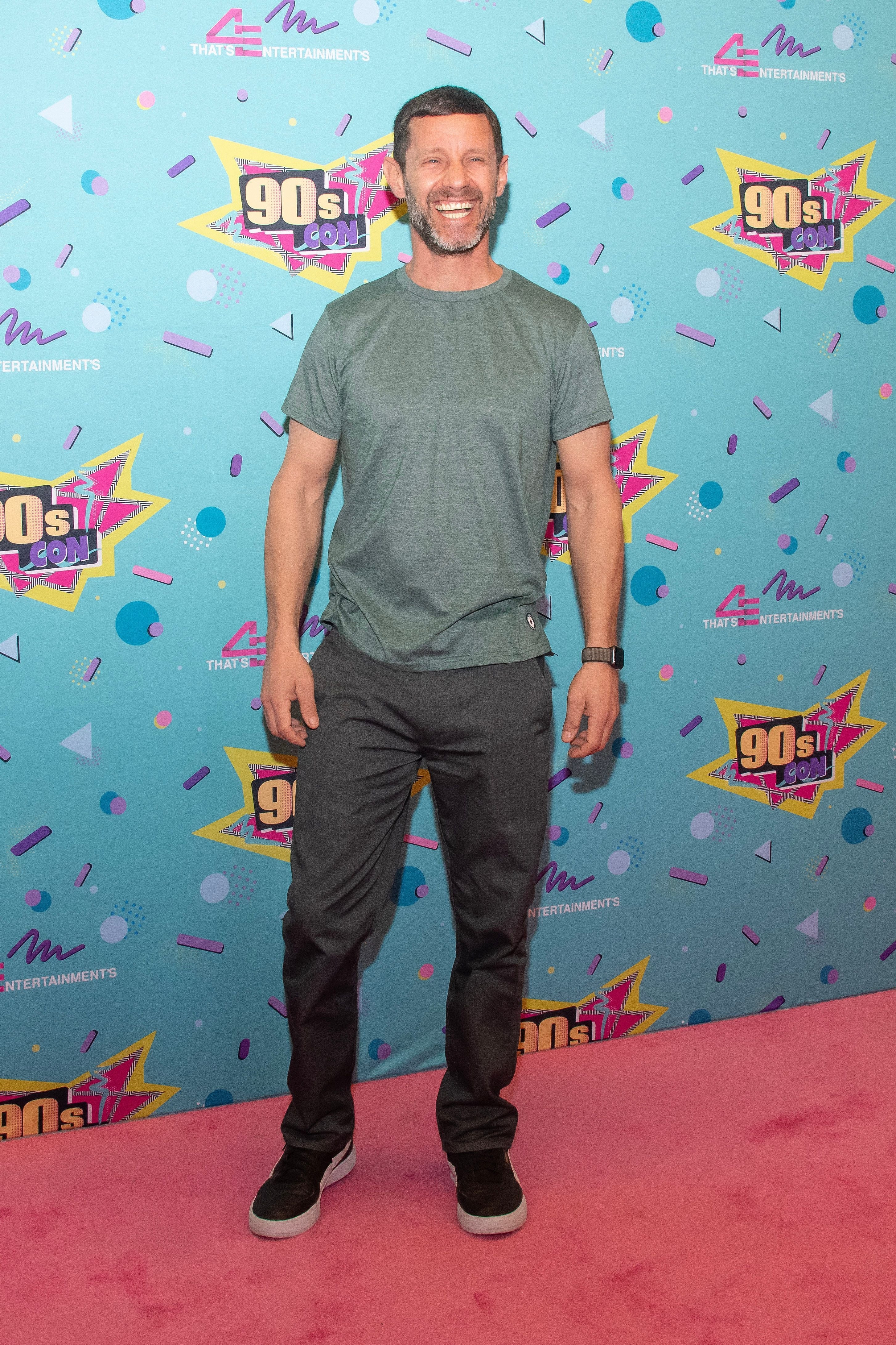 Person smiling on a red carpet, wearing a casual short-sleeve shirt and pants, against a colorful '90s themed background