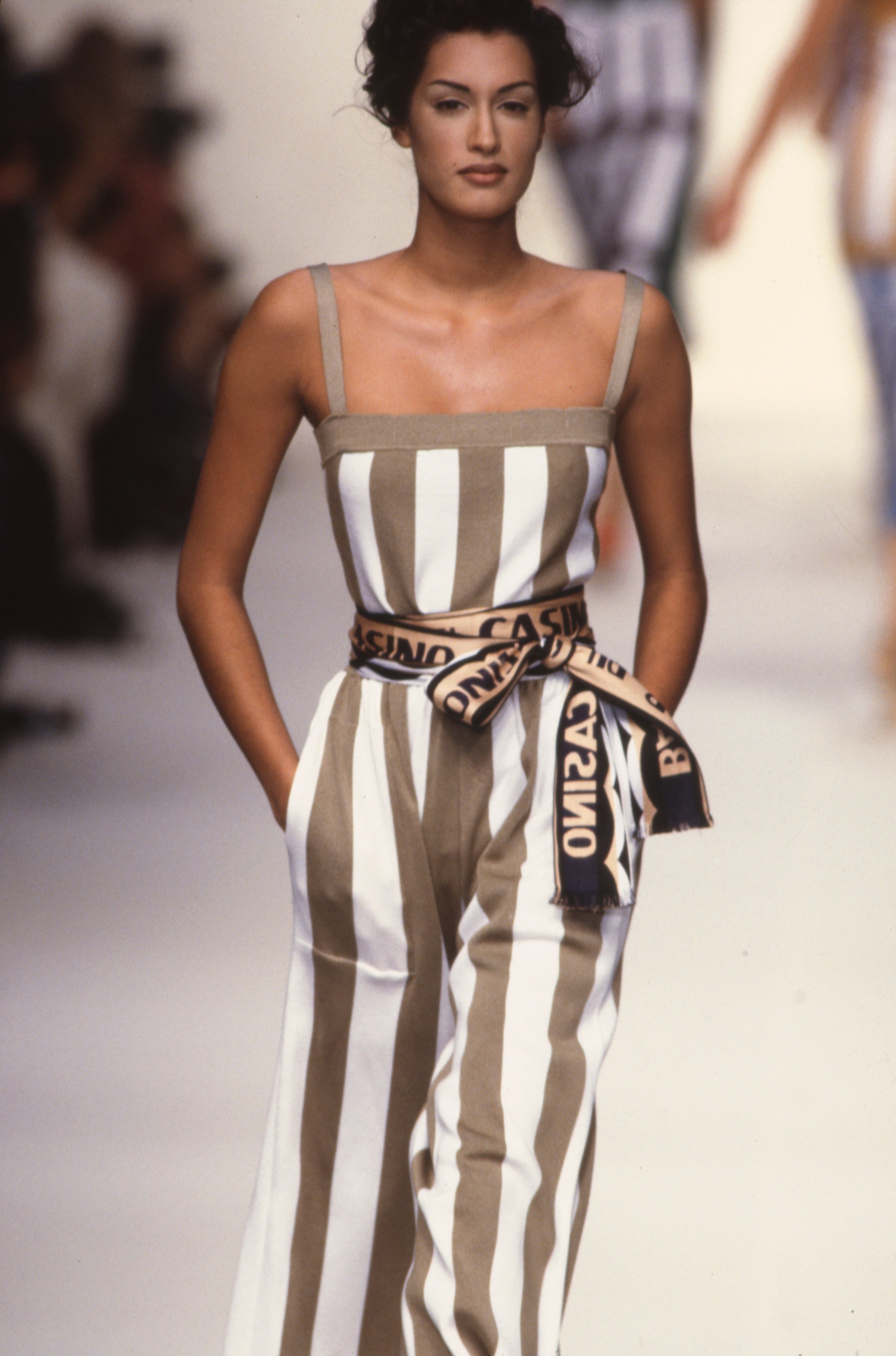 Model on runway wearing a sleeveless, striped jumpsuit with a wide belt, walking confidently