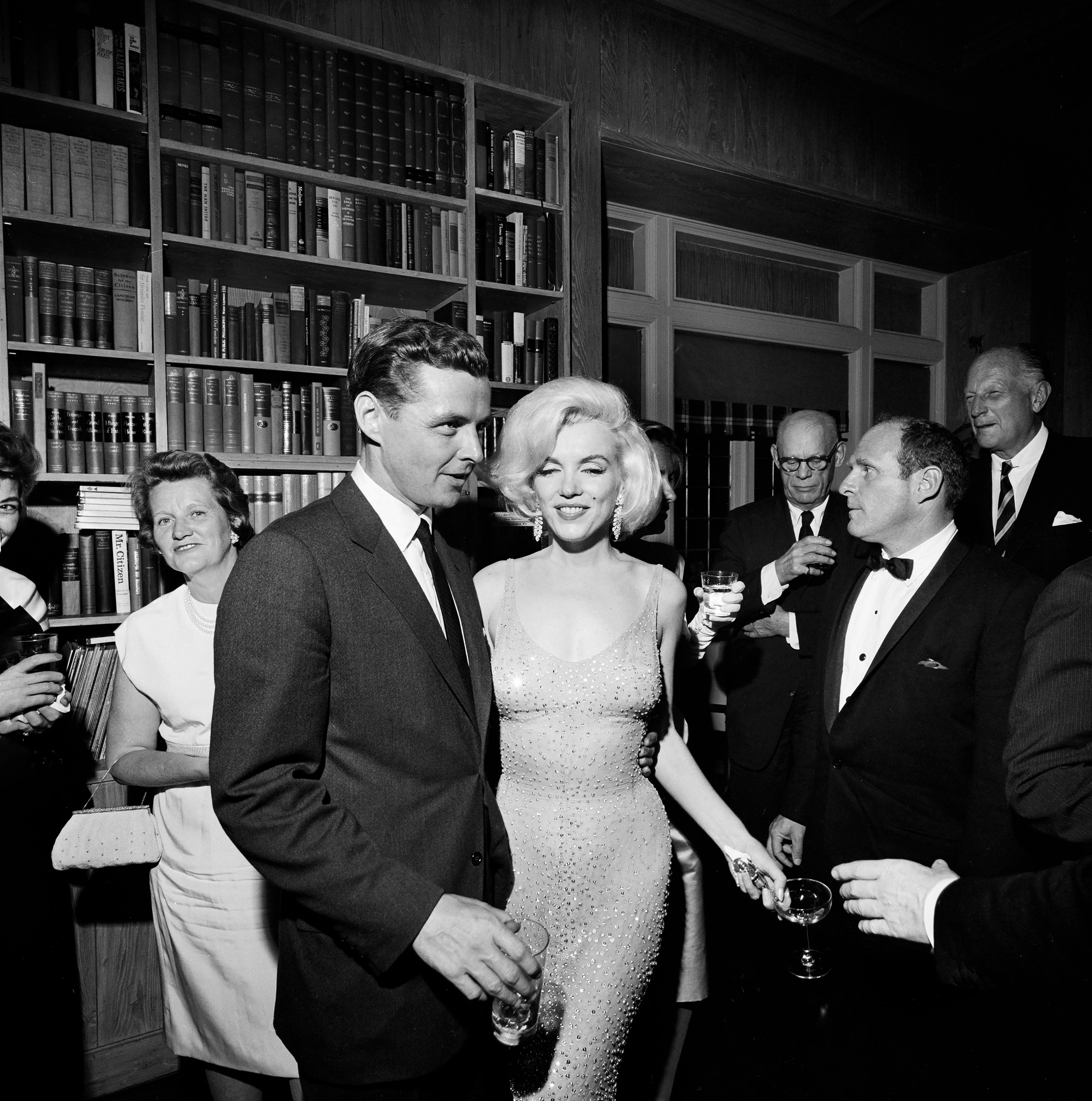 A glamorous Hollywood prima successful a sparkling formal poses astatine an elegant societal gathering with guests successful ceremonial attire, surrounded by bookshelves