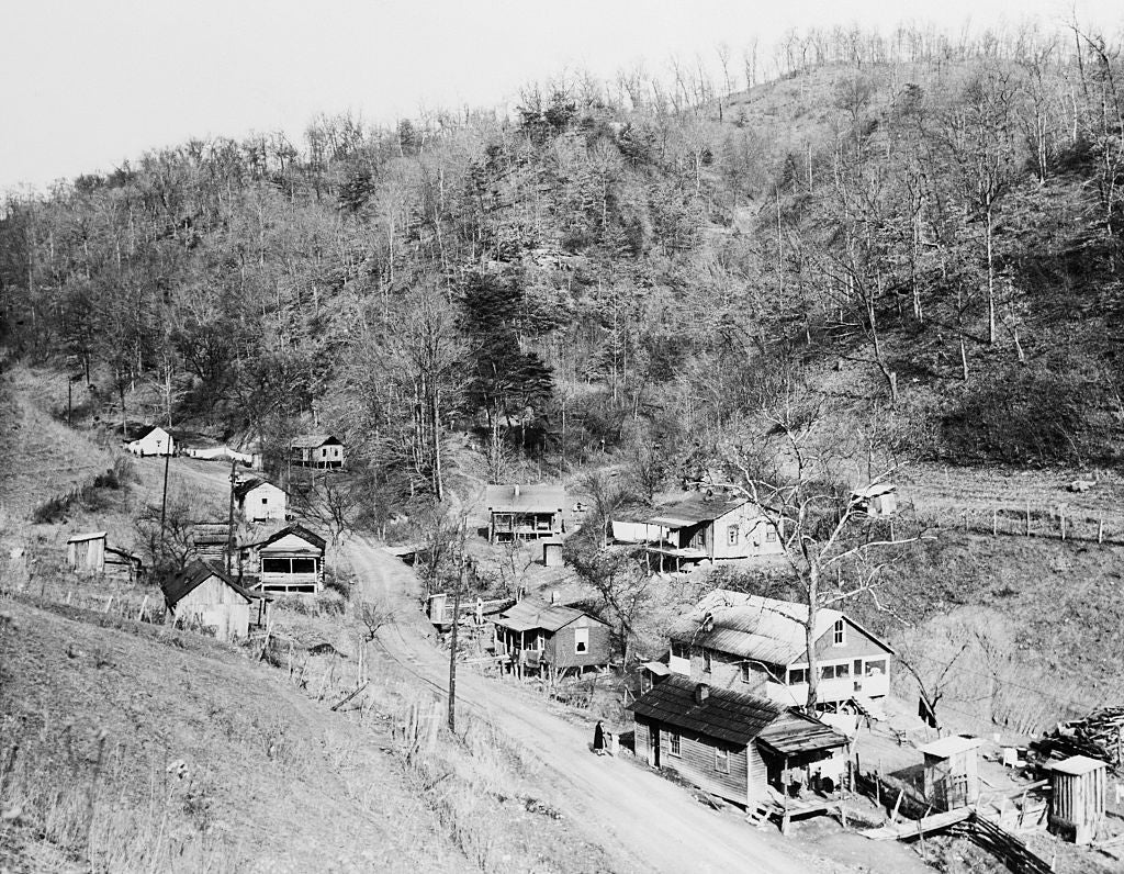 Hillside colony with a ungraded road, tiny woody houses, and sparse trees, reflecting a agrarian mid-20th-century American setting