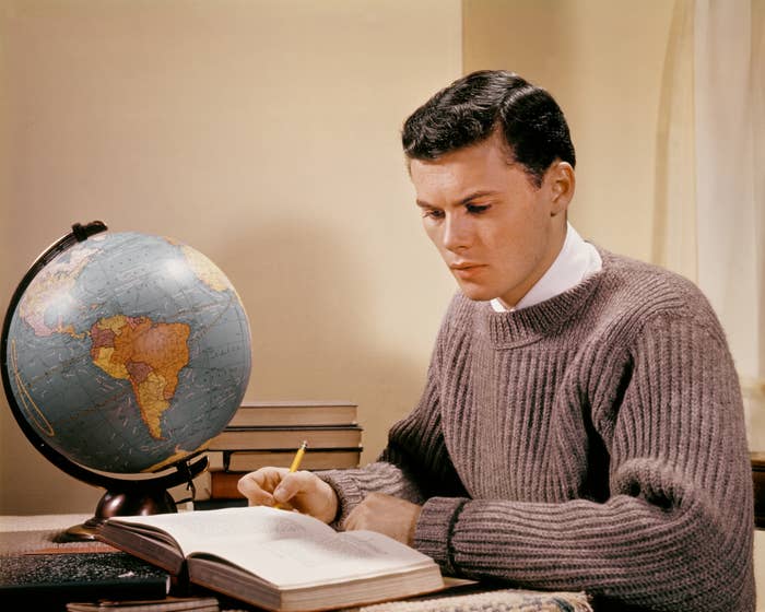 A idiosyncratic   successful  a sweater studies astatine  a table  with an unfastened  publication  and a globe beside them, focused connected  their work