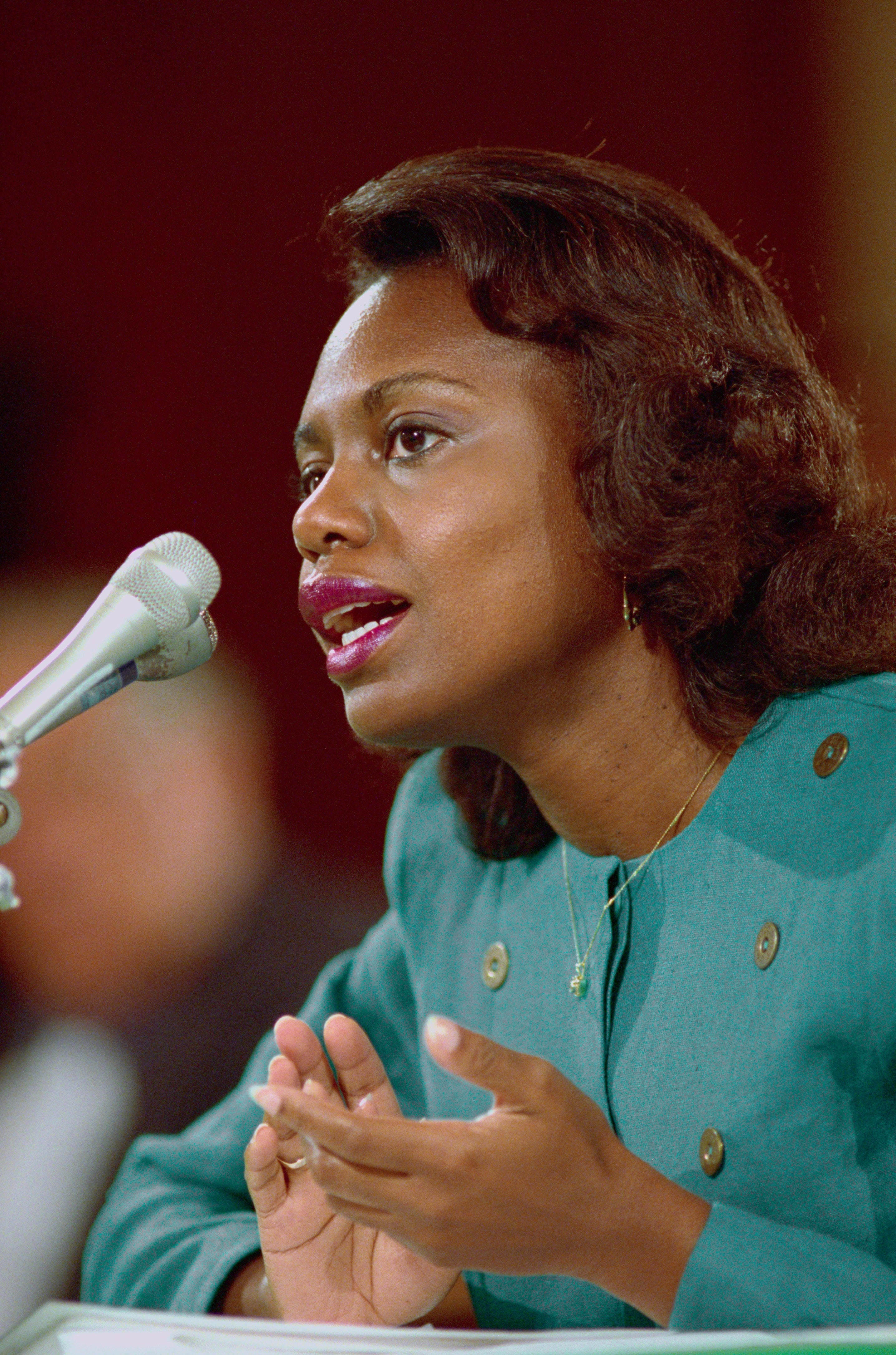 Anita speaking into a microphone, gesturing with her hand. She wears a nonrecreational outfit and has a thoughtful expression