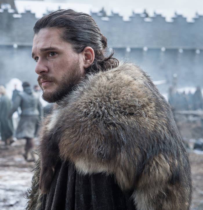 Person in medieval-style fur cloak standing outdoors, looking pensive. Background shows blurred figures and stone walls
