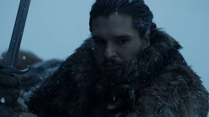 Person in fur cloak holds sword amidst falling snow, looking determined