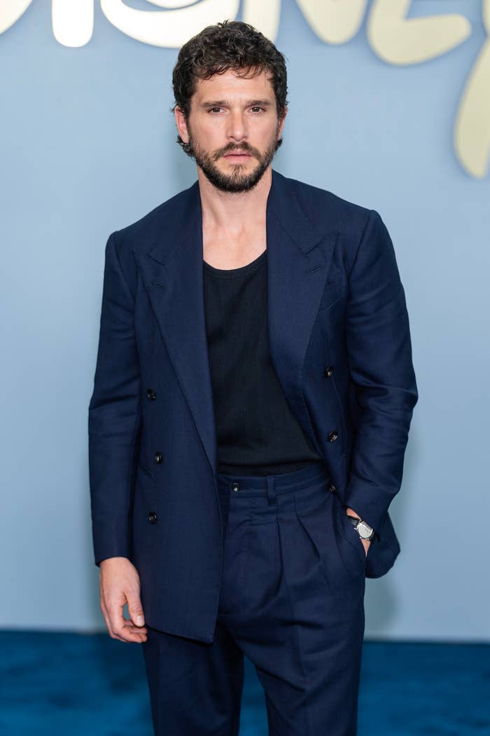 Person in a tailored suit stands confidently on a blue carpet, with a casual black shirt underneath, exuding a modern and stylish look