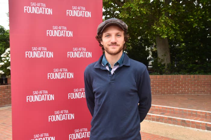 Man in a flat cap and casual sweater posing in front of a SAG-AFTRA Foundation backdrop outdoors