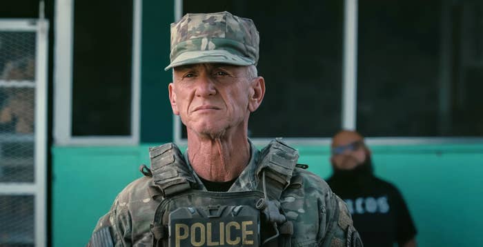 An older man in a military uniform labeled "Police" stands seriously, with another person blurred in the background