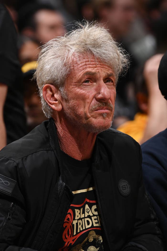 Person sitting in a crowd, wearing a graphic t-shirt and jacket, looking contemplative