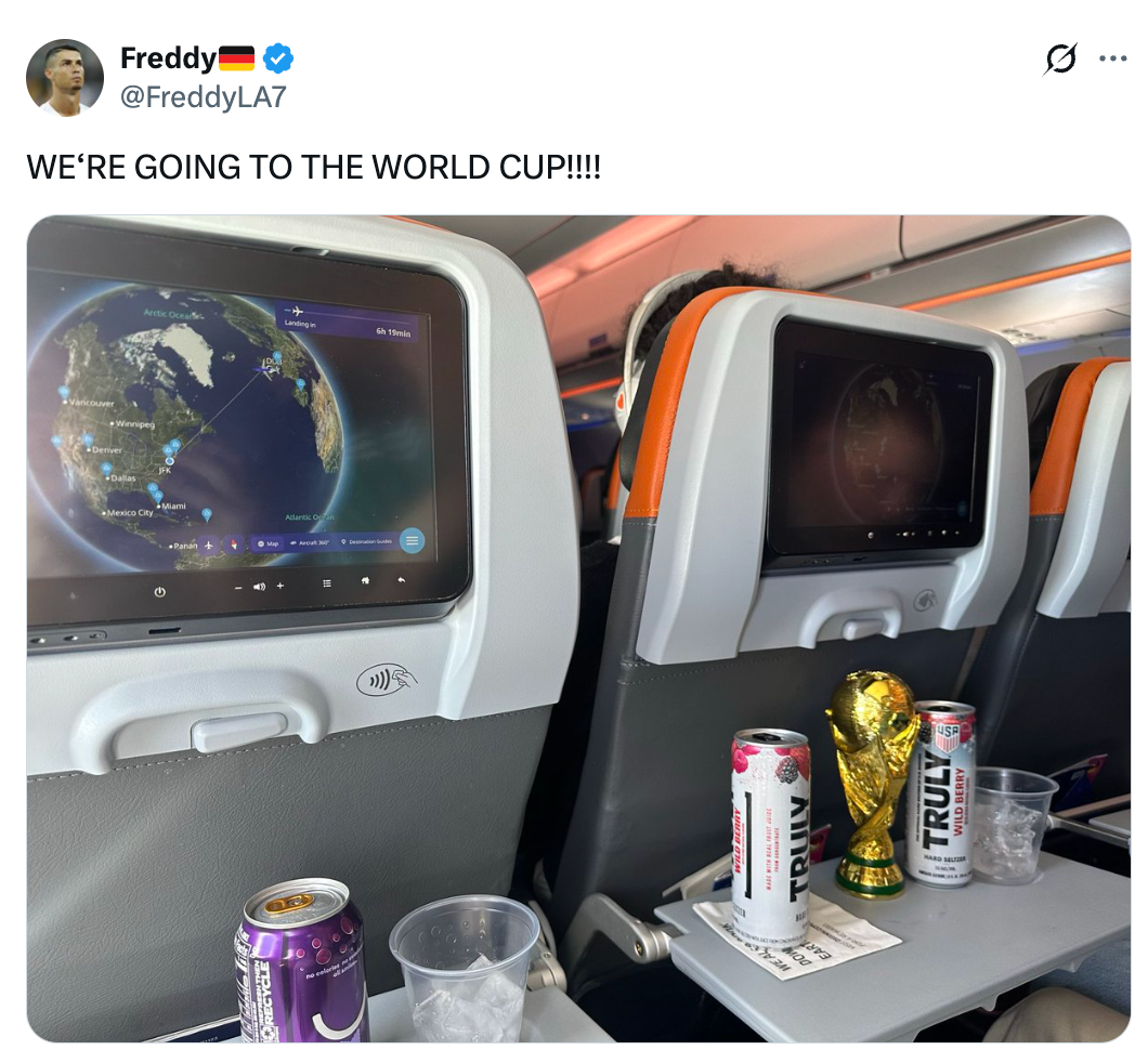 Airplane seatbacks with screens showing a formation map. A tiny World Cup trophy and portion cans are connected the tray table