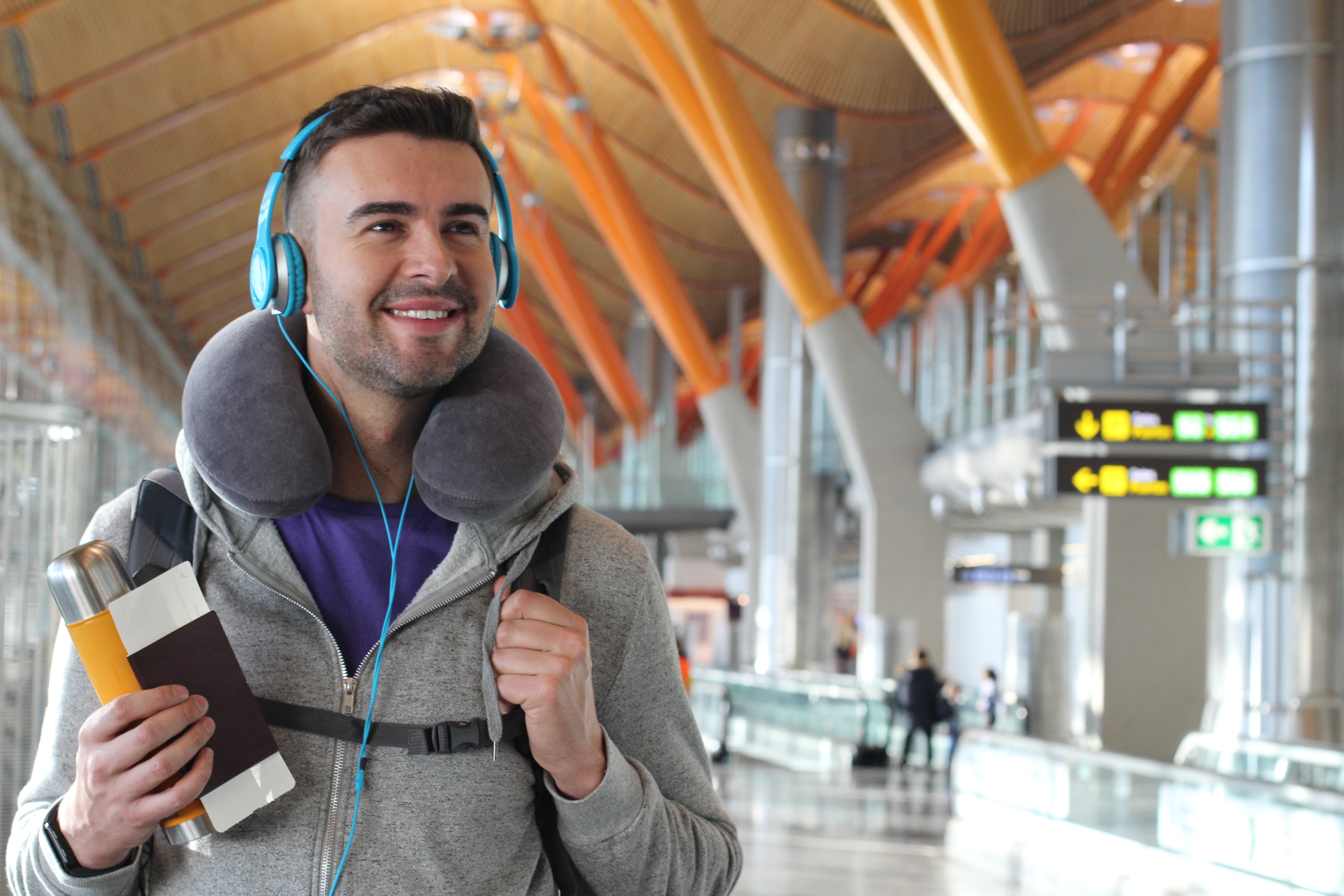 Man at airport smiling, wearing headphones and neck pillow, holding passport and journal, ready for travel
