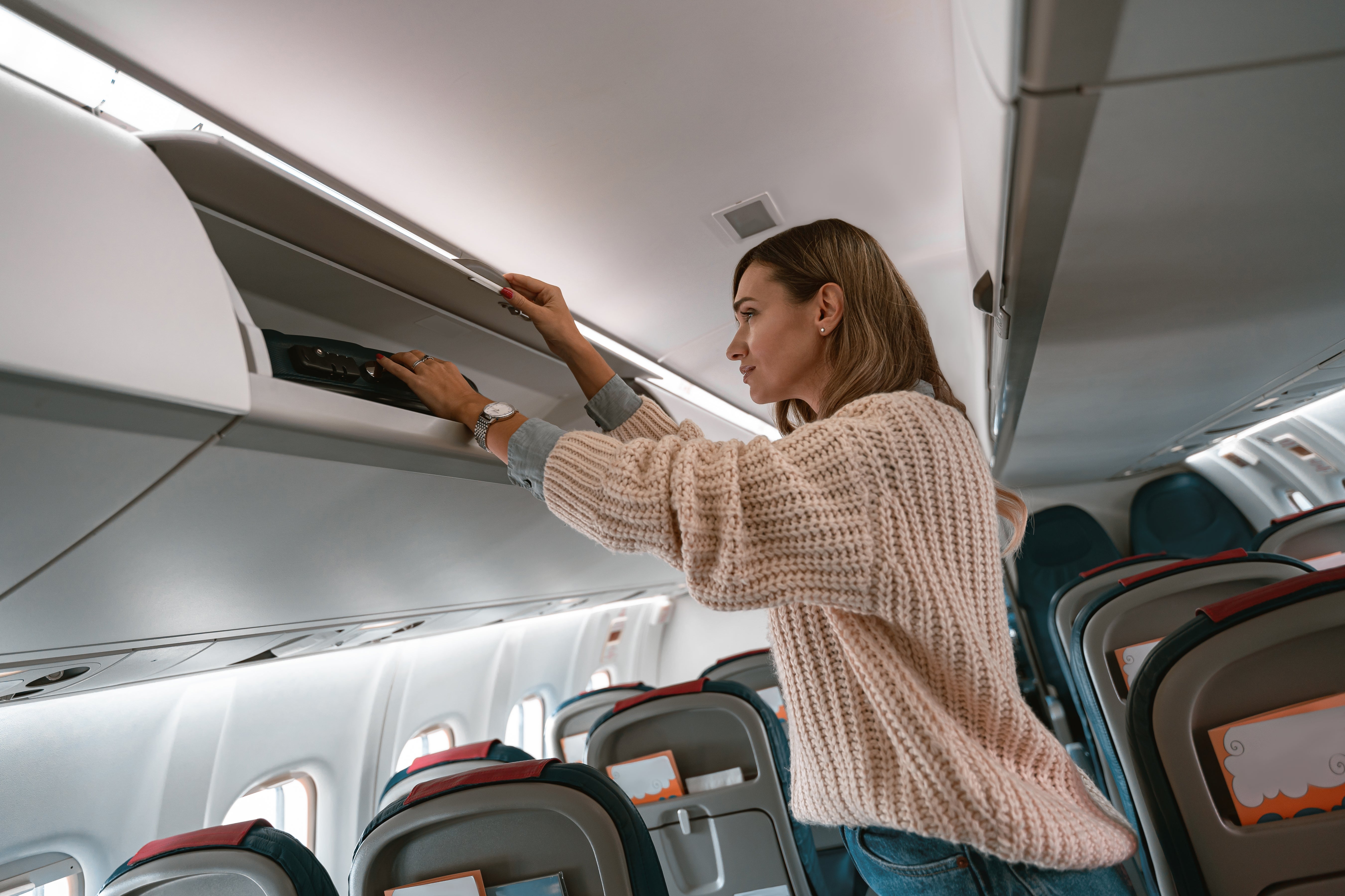 A person in a cozy sweater placing a bag in an airplane's overhead compartment