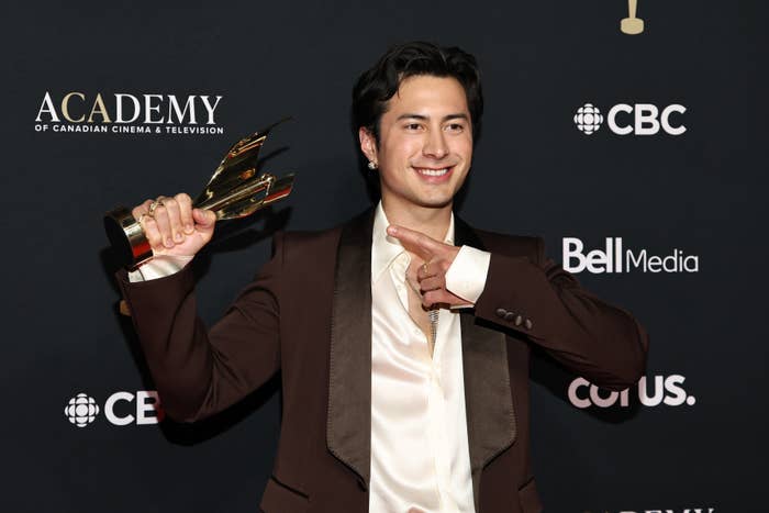 Person in a smart suit holds a trophy on a red carpet, smiling and pointing. Background shows event sponsor logos
