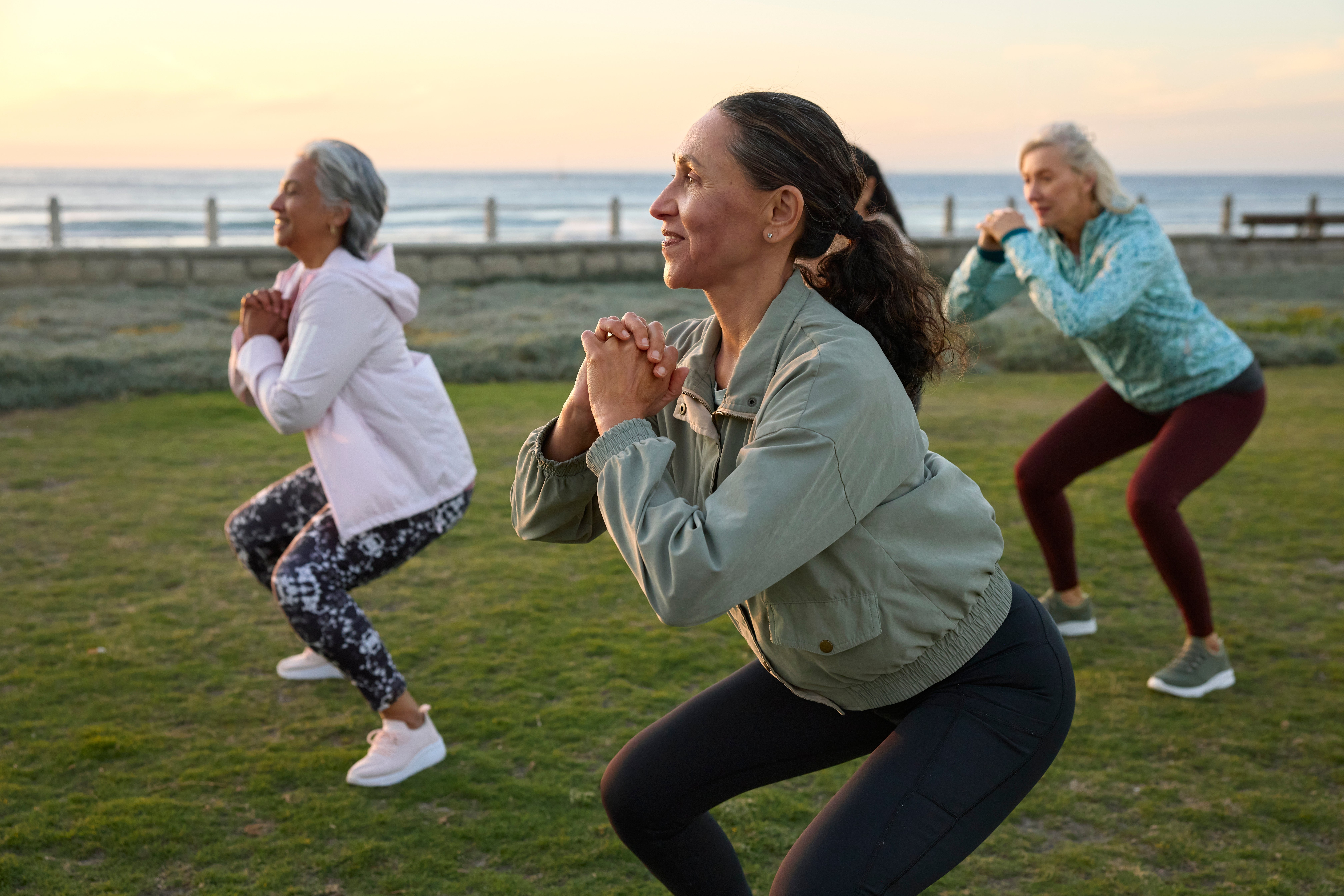 Three women exercising outdoors by the water, performing squats unneurotic connected a grassy area