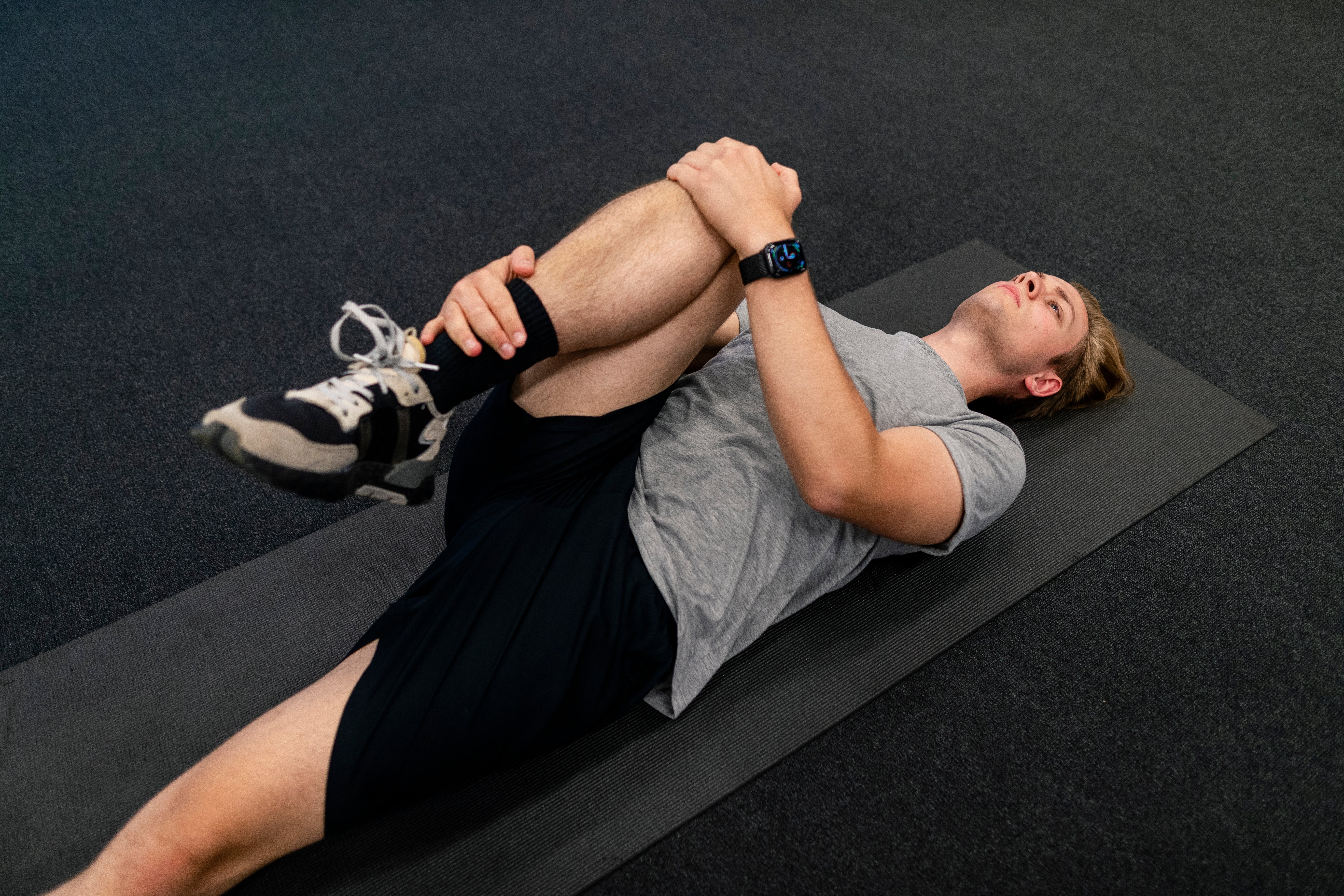Person lying connected a mat, stretching 1 limb and holding it with some hands, wearing diversion covering and a smartwatch