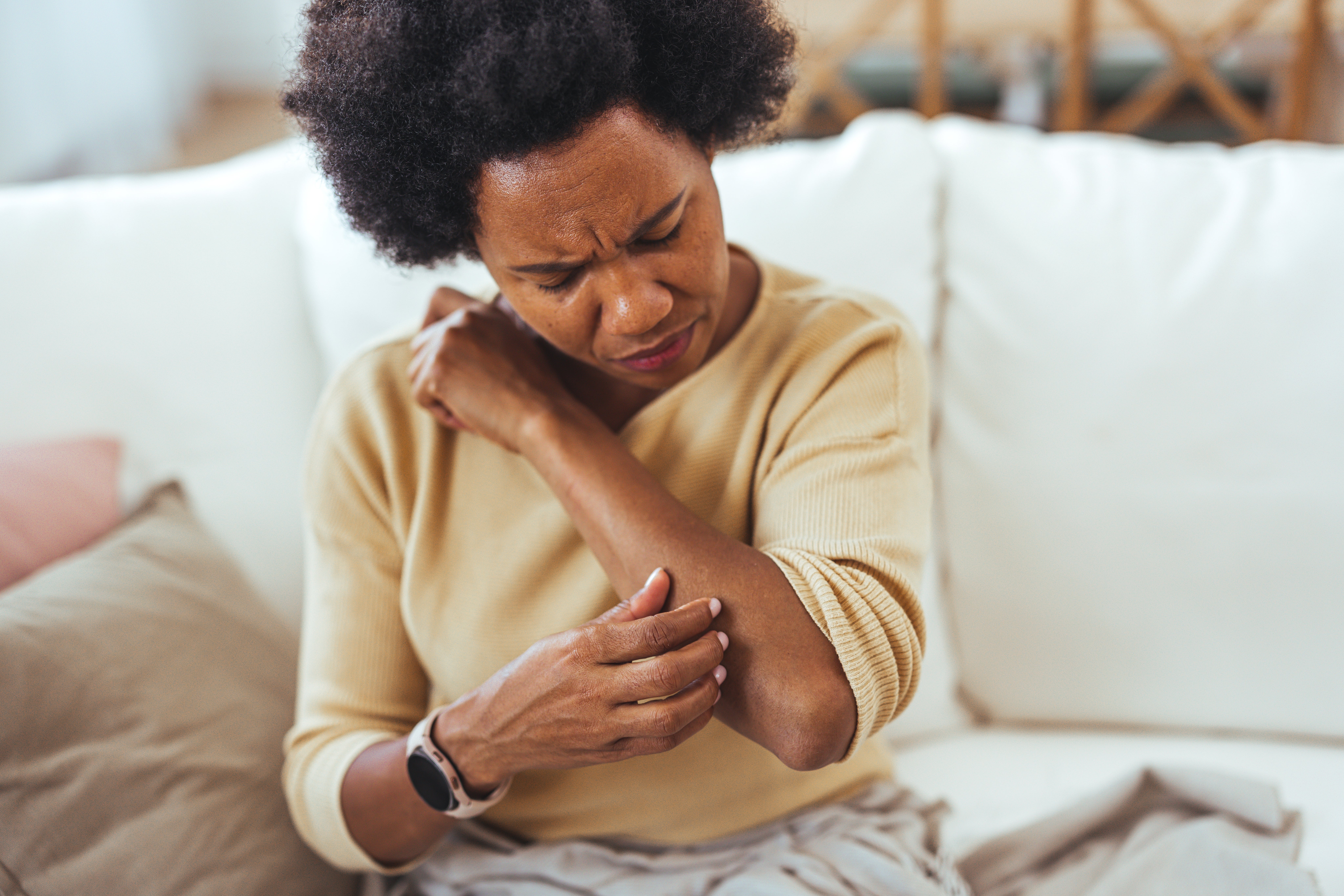 Woman sitting connected  a sofa  scratches her elbow, appearing uncomfortable. She wears a astute  ticker  and a casual long-sleeved shirt