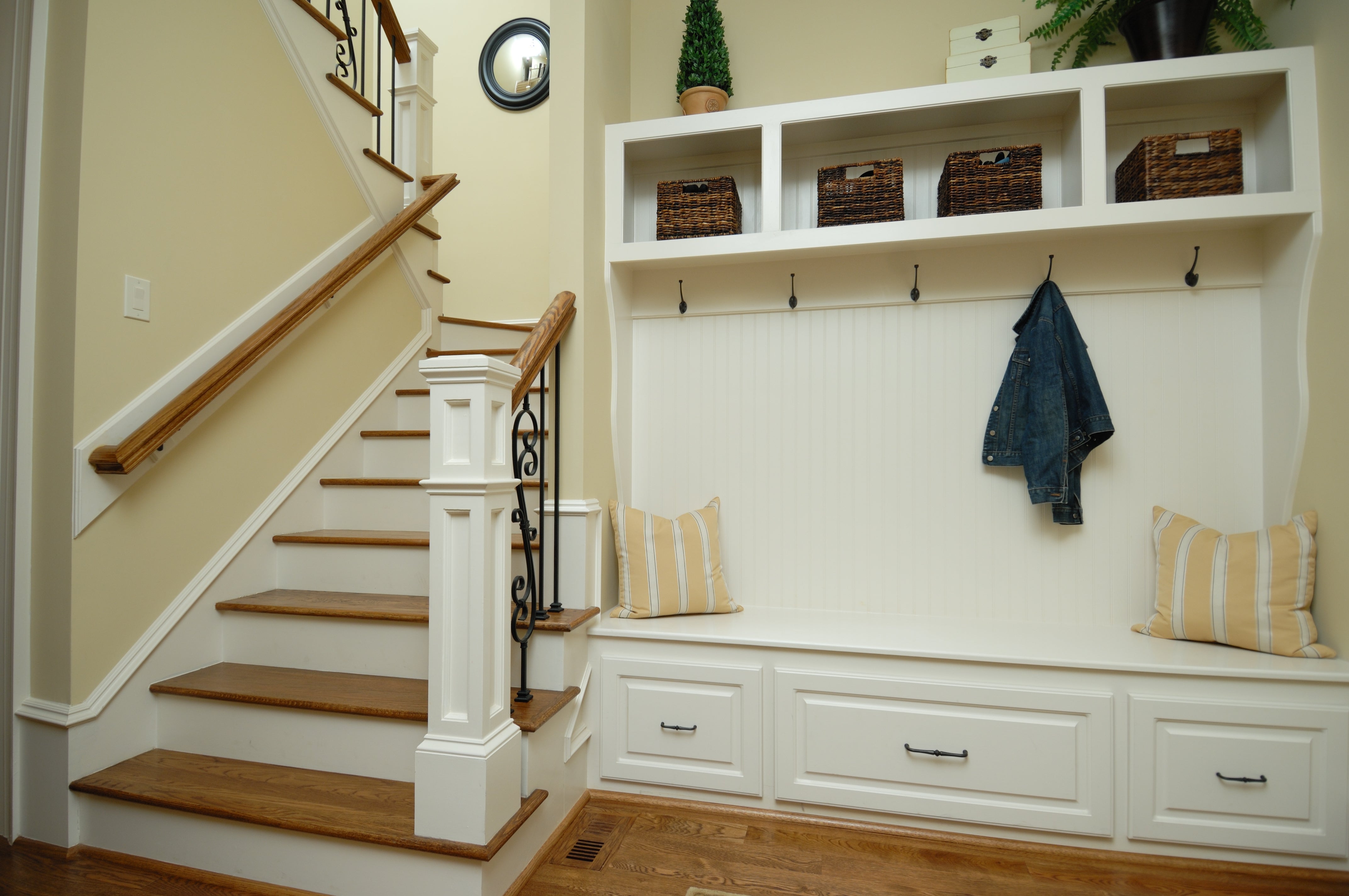 Entryway with woody staircase, achromatic built-in seat with drawers, baskets connected shelf, hooks, and a denim overgarment hanging. Cozy seating with pillows