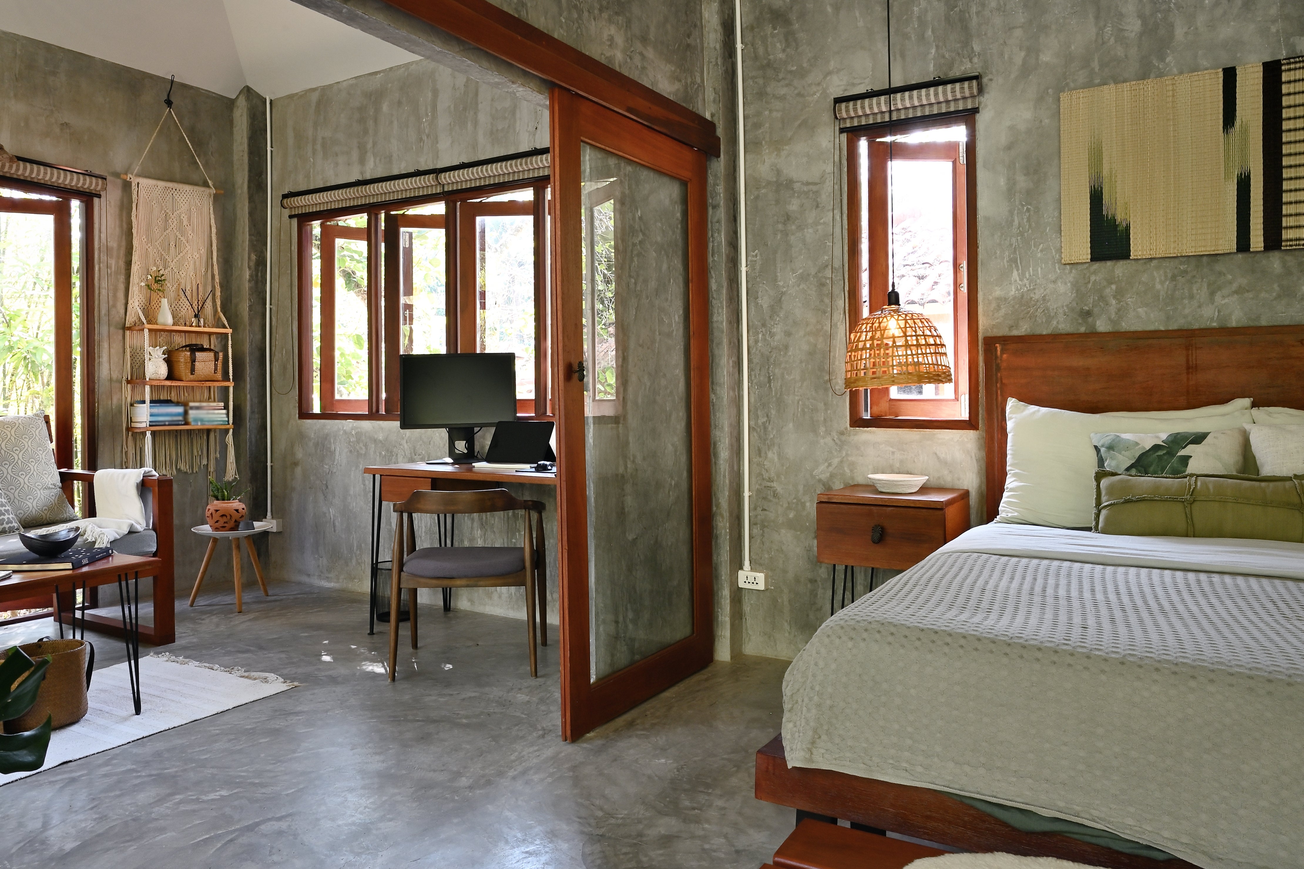 Modern chamber and workspace with factual walls, woody accents, table with computer, and a bed. Large windows supply earthy light