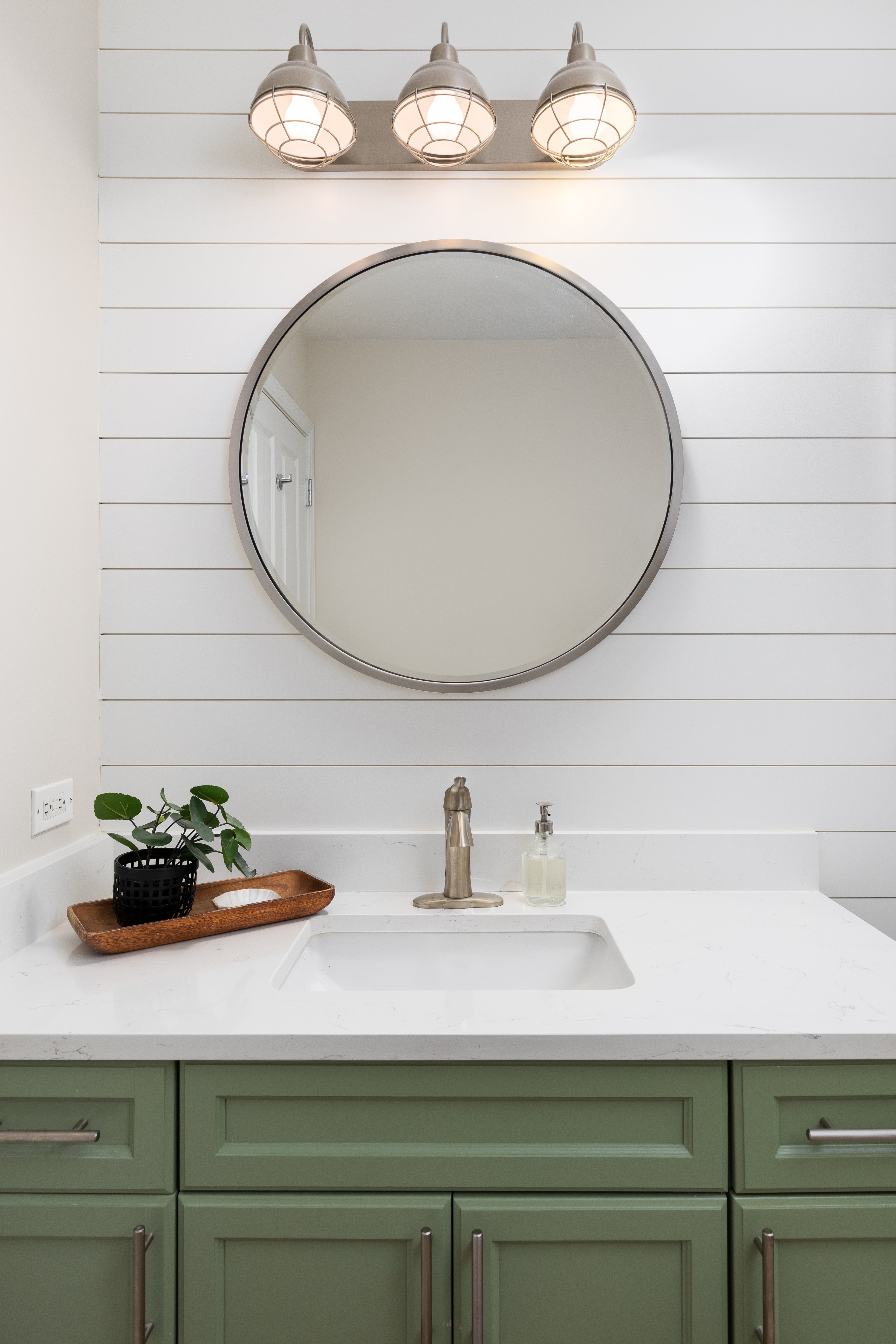Bathroom vanity with circular mirror, achromatic countertop, greenish cabinets, and wall-mounted lights. A tiny works and soap dispenser are connected the counter