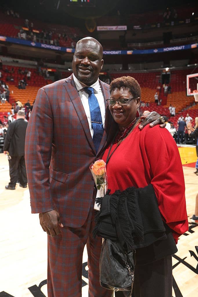 A tall man in a plaid suit with a blue tie stands smiling with an older woman holding a bouquet on an indoor basketball court