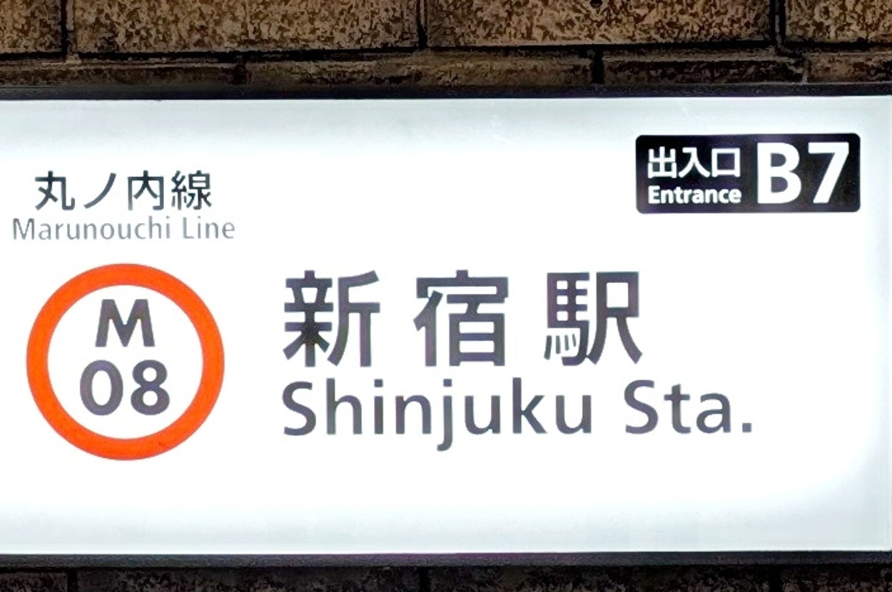 新宿駅の看板がある階段口で、下側には飲食店のメニューが並んでいる。