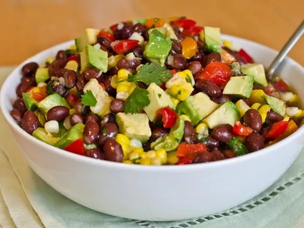Bowl of mixed bean salad with avocado, corn, bell peppers, and cilantro, sitting on a cloth with a spoon in the bowl