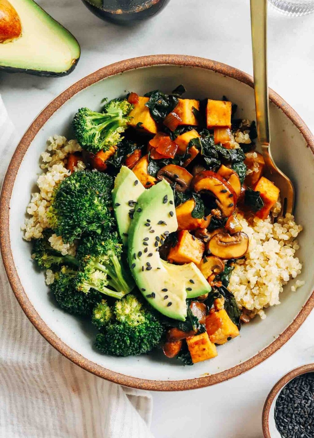 A bowl of quinoa topped with roasted sweet potatoes, broccoli, avocado slices, and sesame seeds