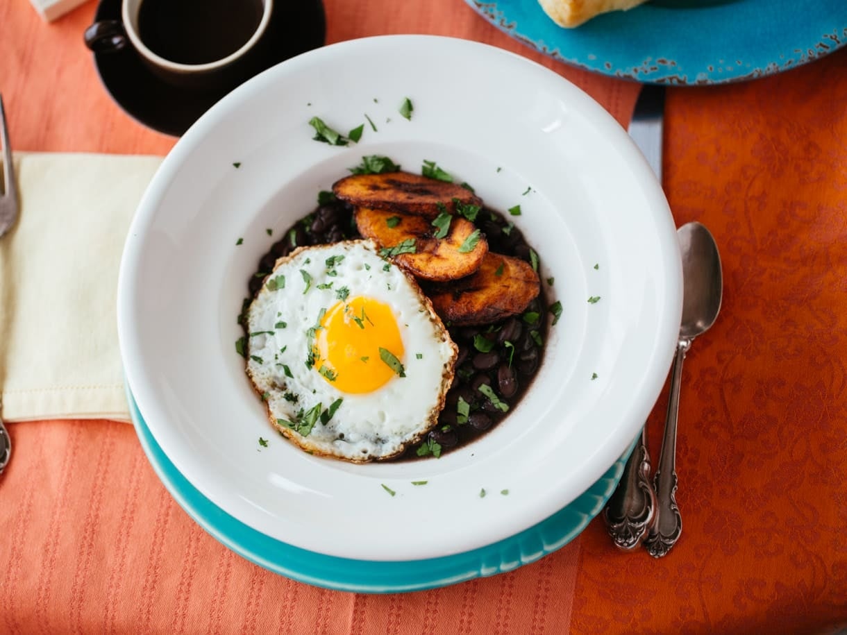 A plate with a sunny-side-up egg, fried plantains, and black beans, garnished with herbs. A cup of coffee is in the background