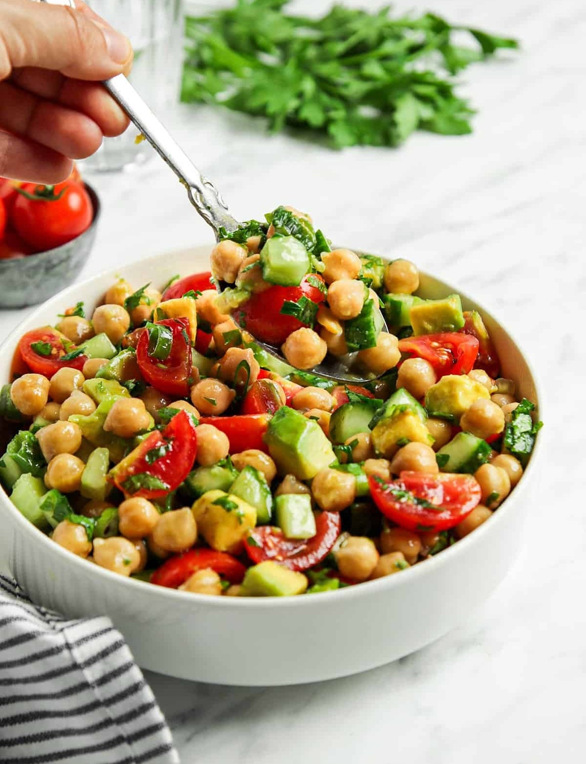 A hand holding a fork over a bowl of chickpea salad with tomatoes, cucumber, and herbs on a marble surface