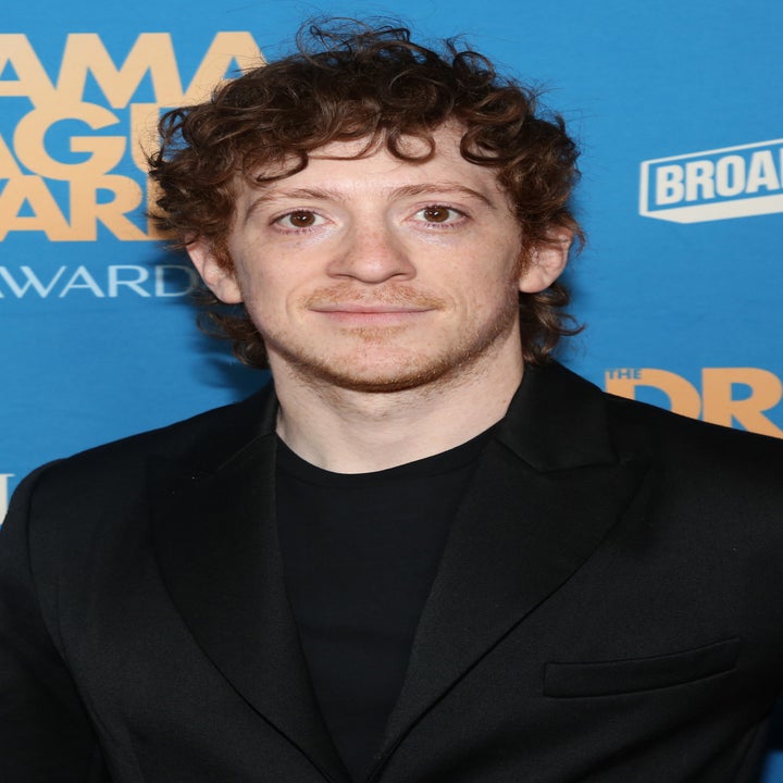 A person with curly hair in a black suit poses at an event with a blue backdrop featuring various logos and award names