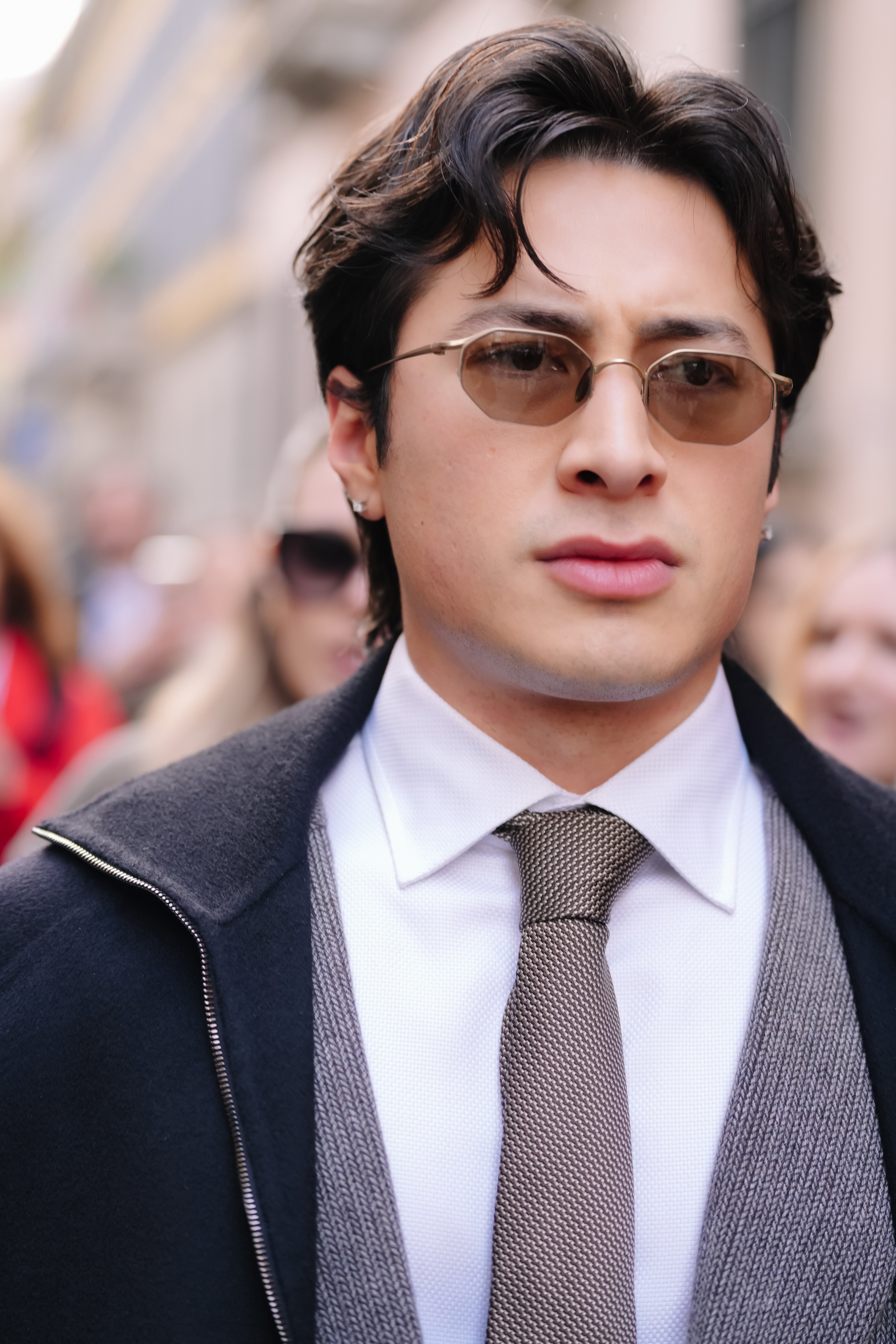 Person wearing a suit with a tie over a shirt and a coat. They have short hair and sunglasses, walking in a bustling urban setting