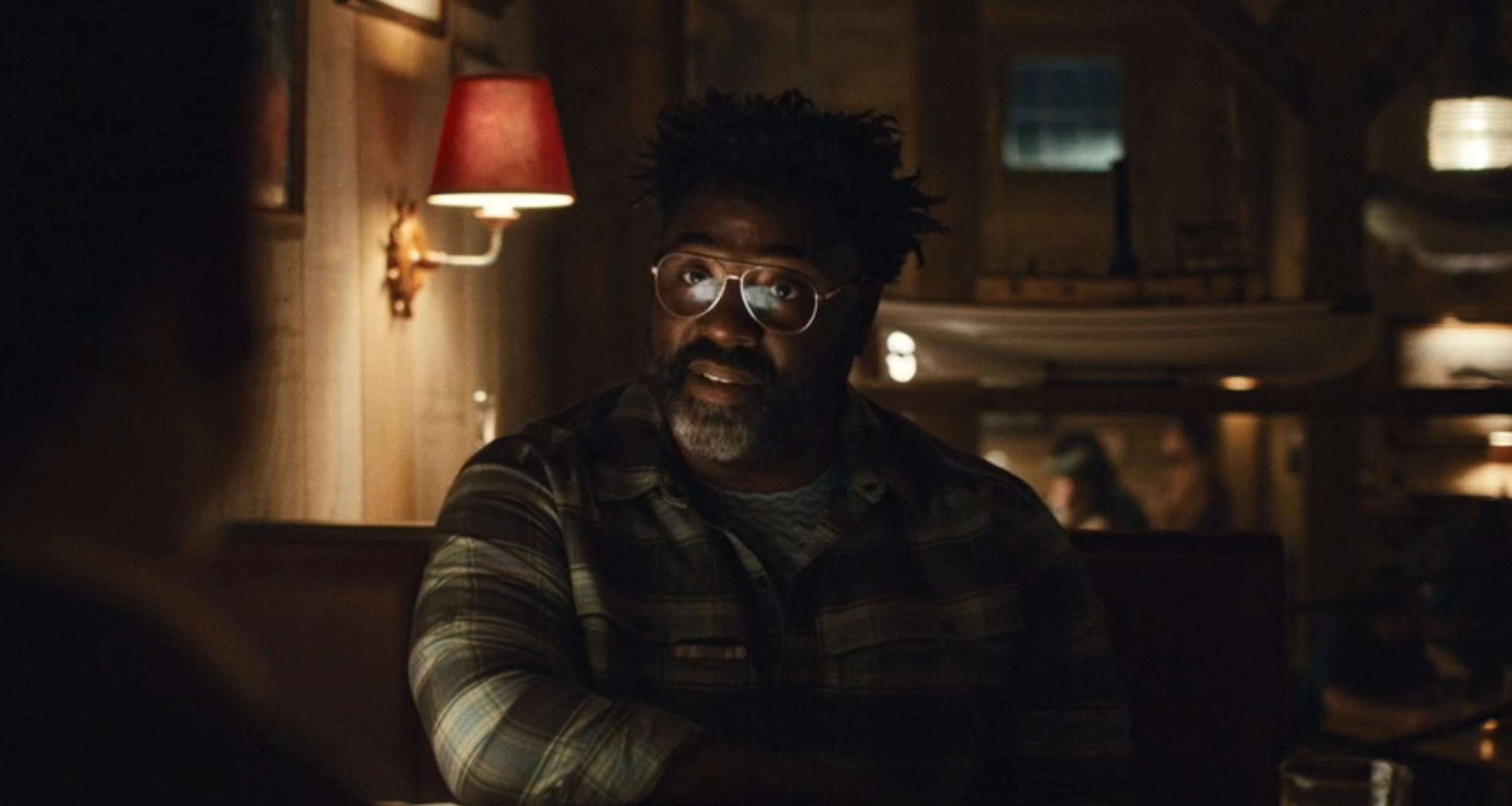 A antheral   with glasses and a plaid garment  sits astatine  a dimly lit edifice  booth, engaged successful  conversation