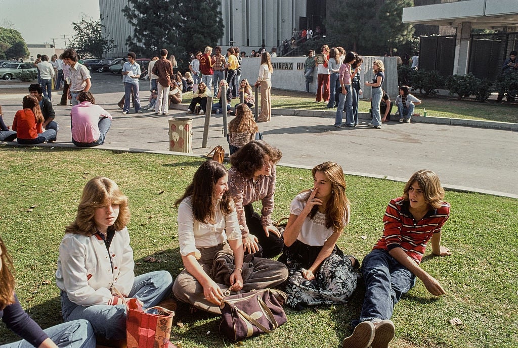 Remaja duduk di rumput, berbicara dan bersantai di dekat gedung sekolah. Para siswa mengenakan pakaian kasual bergaya tahun 1970-an, menikmati hari yang cerah