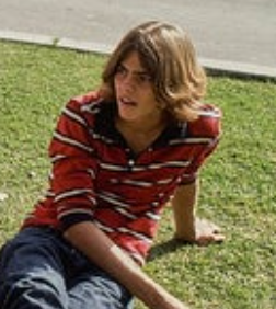 Seseorang dengan rambut sebahu duduk santai di atas rumput, mengenakan kemeja bergaris dan celana jeans, tampak santai dan penuh perhatian