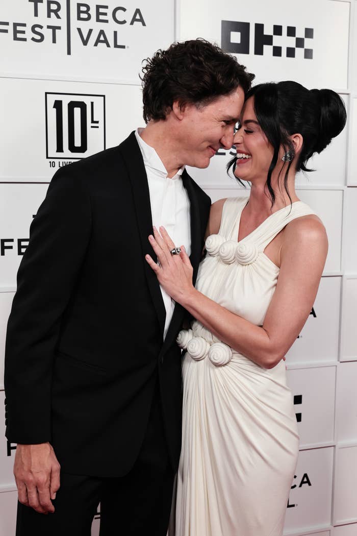 Two people on a red carpet, smiling and embracing. One wears a classic suit, the other a sleeveless gown with textured embellishments