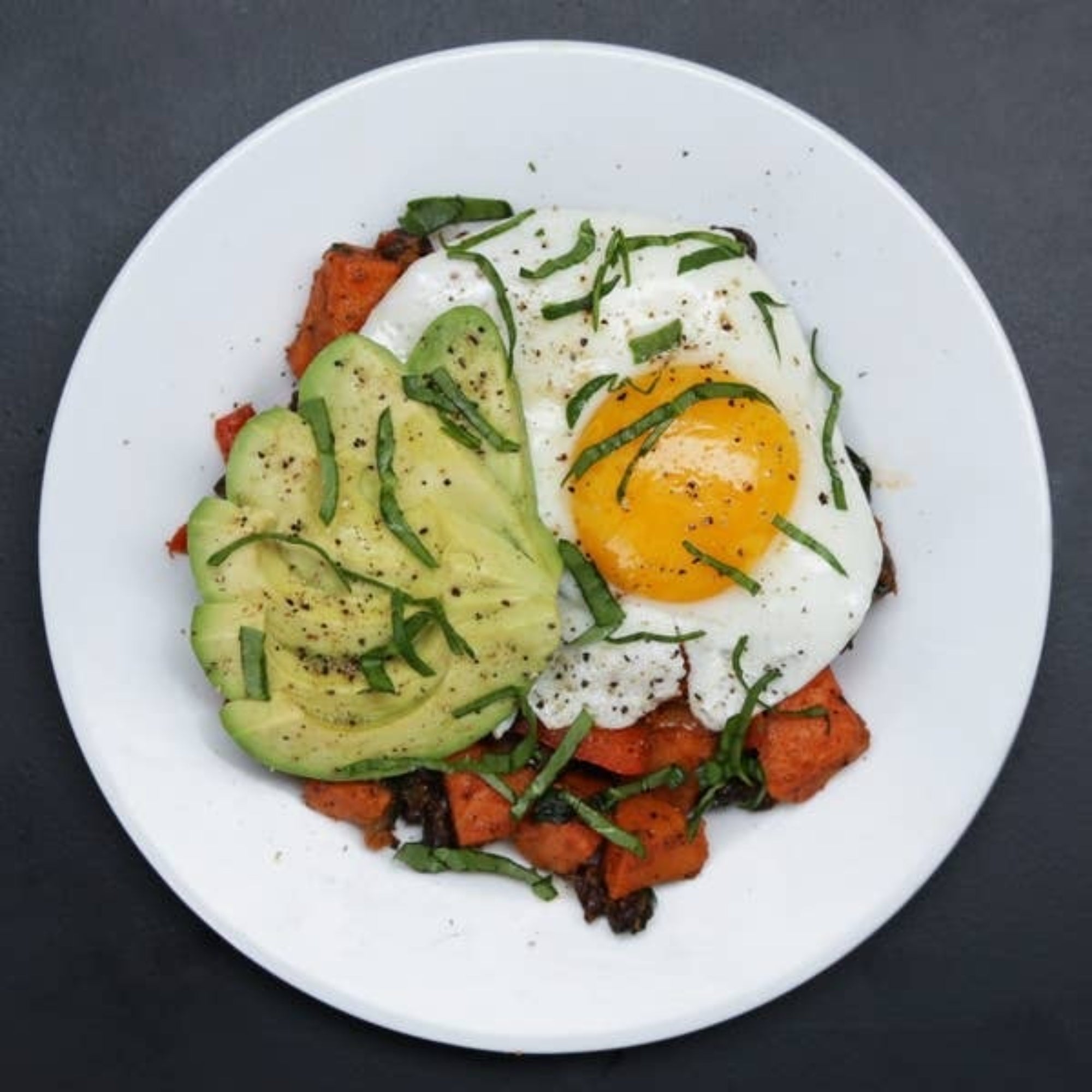 A plate with sweet potatoes, avocado slices, a sunny-side-up egg, garnished with herbs