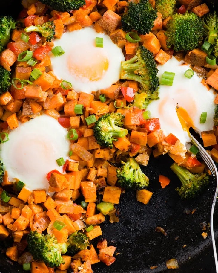 Skillet breakfast with eggs, diced sweet potatoes, broccoli, and peppers. One egg is broken with yolk running