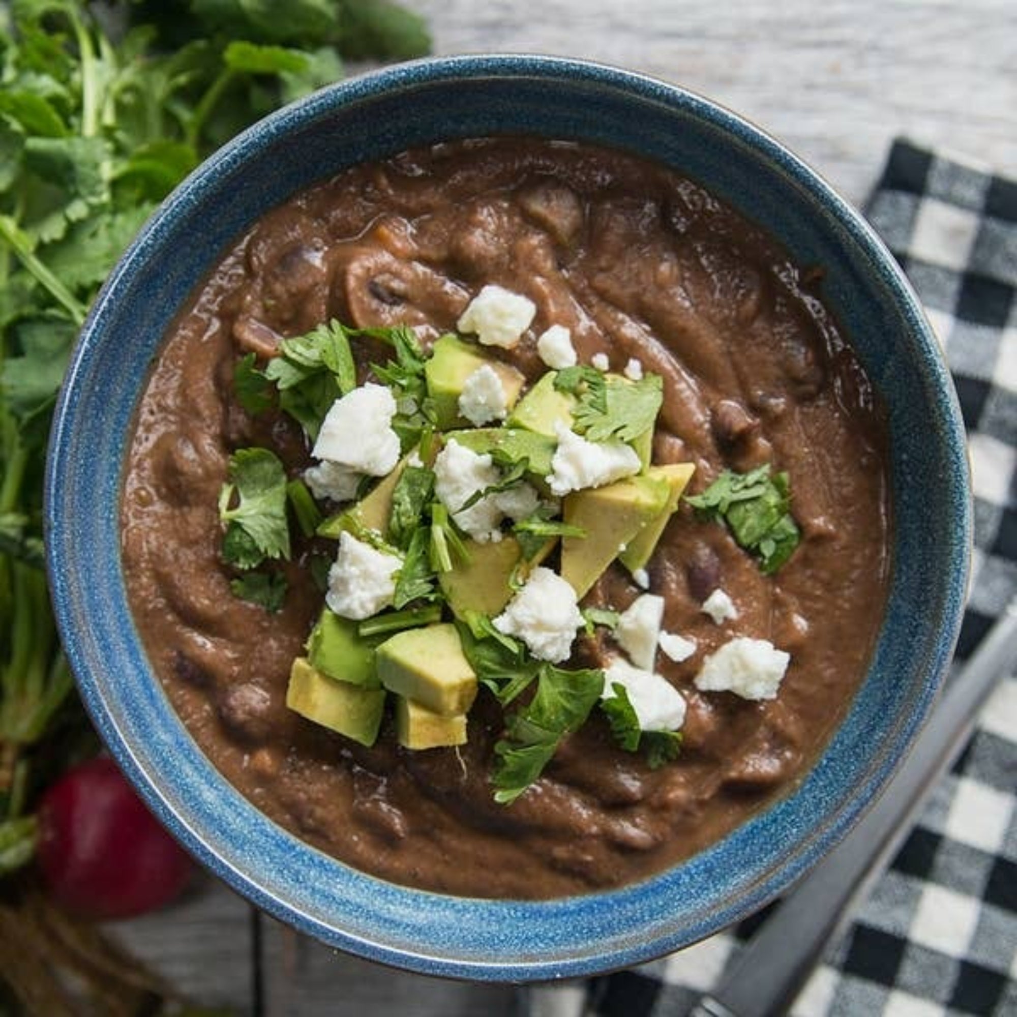 A bowl of black bean soup topped with avocado, cilantro, and crumbled cheese, placed next to a checkered napkin