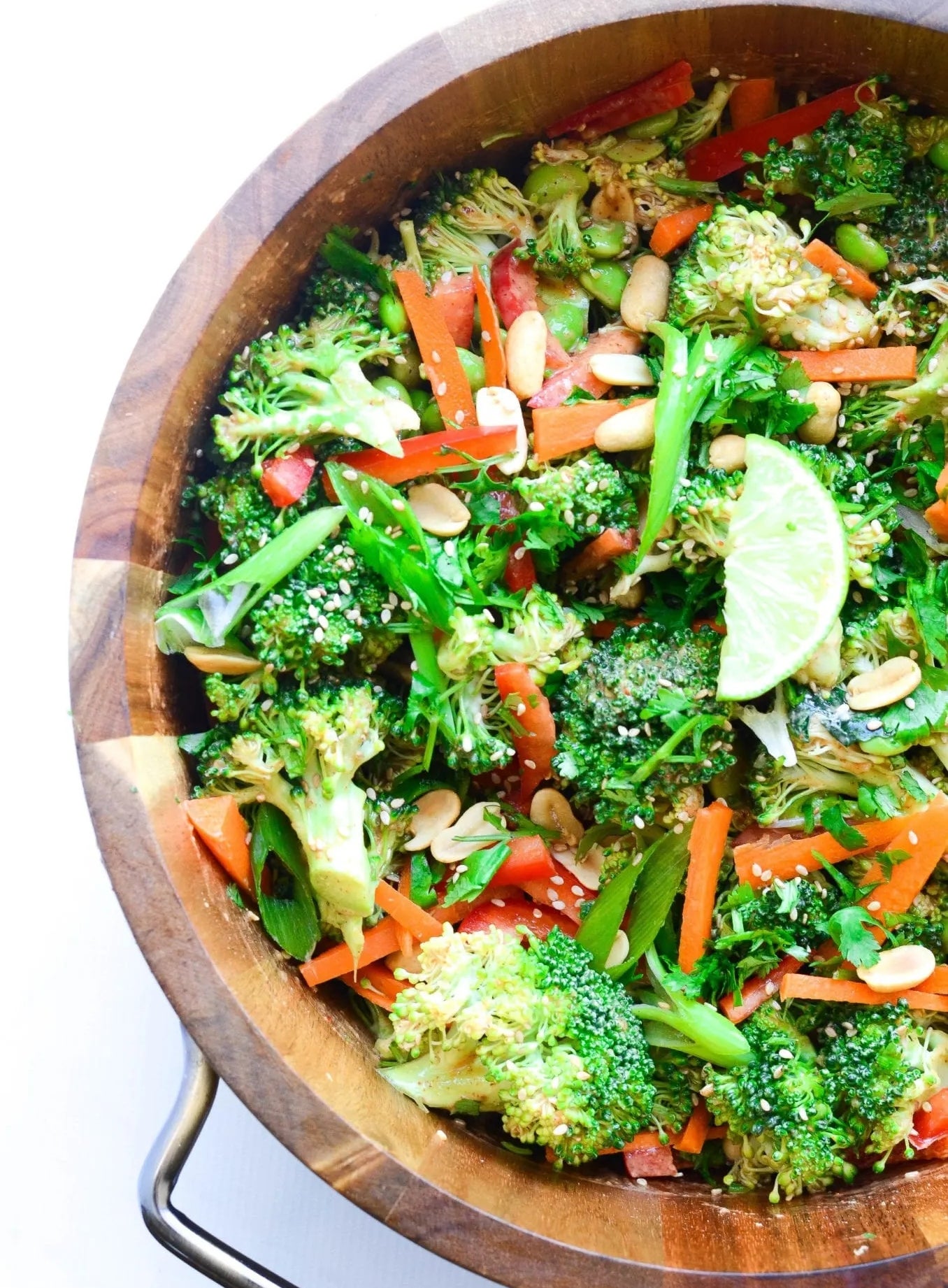 A wooden bowl filled with a broccoli salad, featuring carrots, peanuts, green onions, sesame seeds, and lime wedges