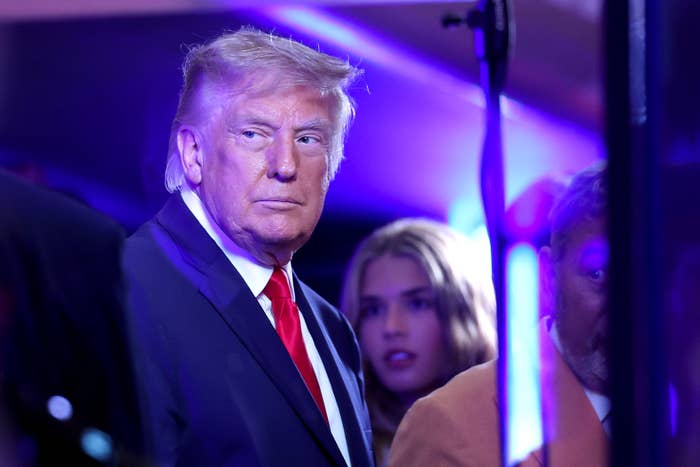 A man in a suit and red tie stands under bright lights, with people in the background