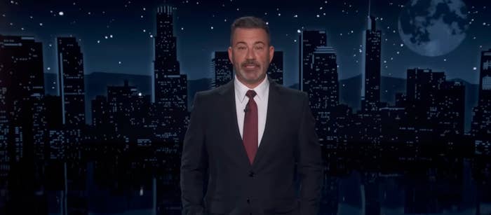 Man in a suit with a tie stands in front of a nighttime city skyline backdrop on a stage