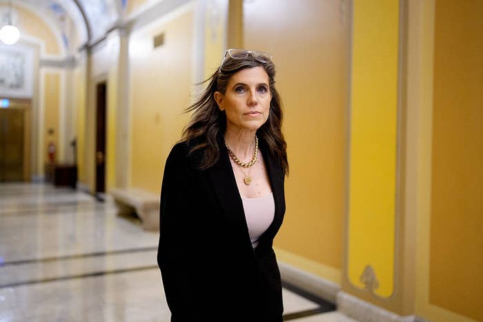 A woman walks confidently through a building hallway, wearing a black blazer and a necklace