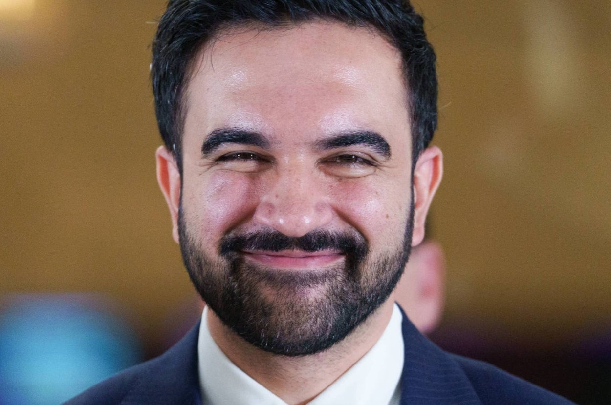A person smiling while wearing a suit and tie, standing in a well-lit environment