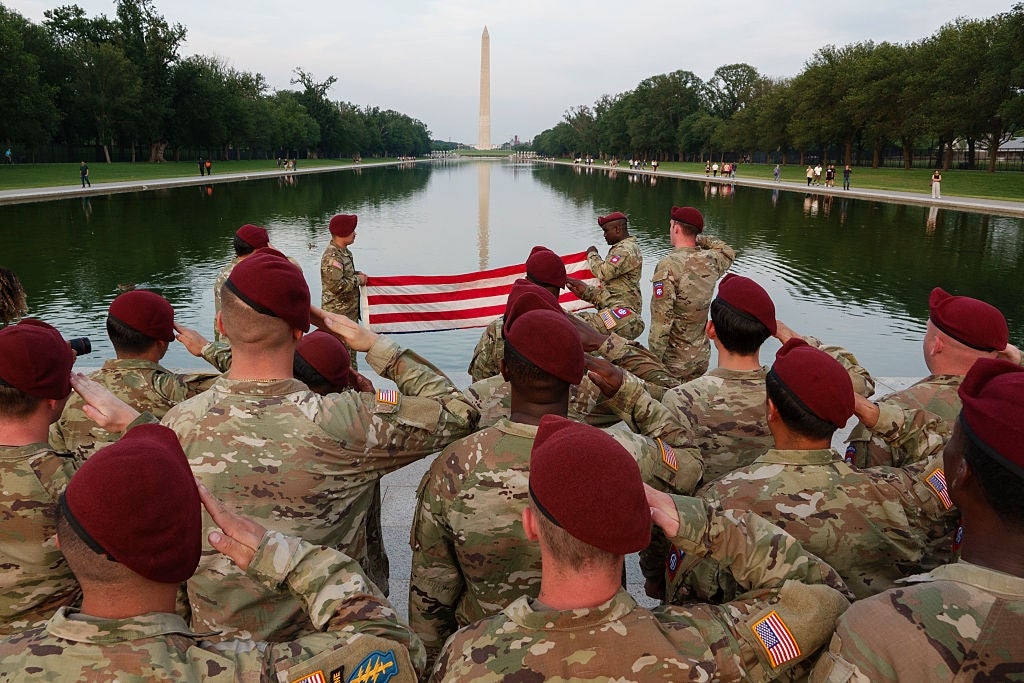 Soldater i uniform og rødbrune basker hilser mens de holder et foldet flagg nær Washington Monument og Reflection Pool