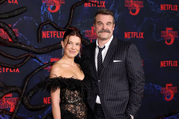Two radical   connected  a reddish  carpet; 1  successful  a achromatic  feathered gown and the different   successful  a striped suit. Netflix and "Stranger Things" logos successful  the background