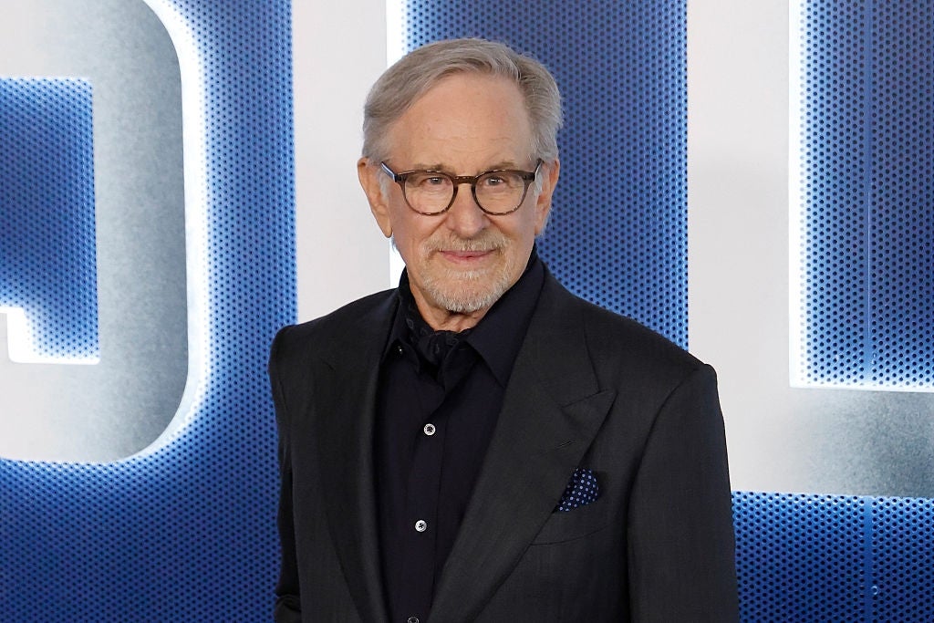 A man in a dark suit and glasses poses confidently at an event, standing in front of a backdrop with large lettering
